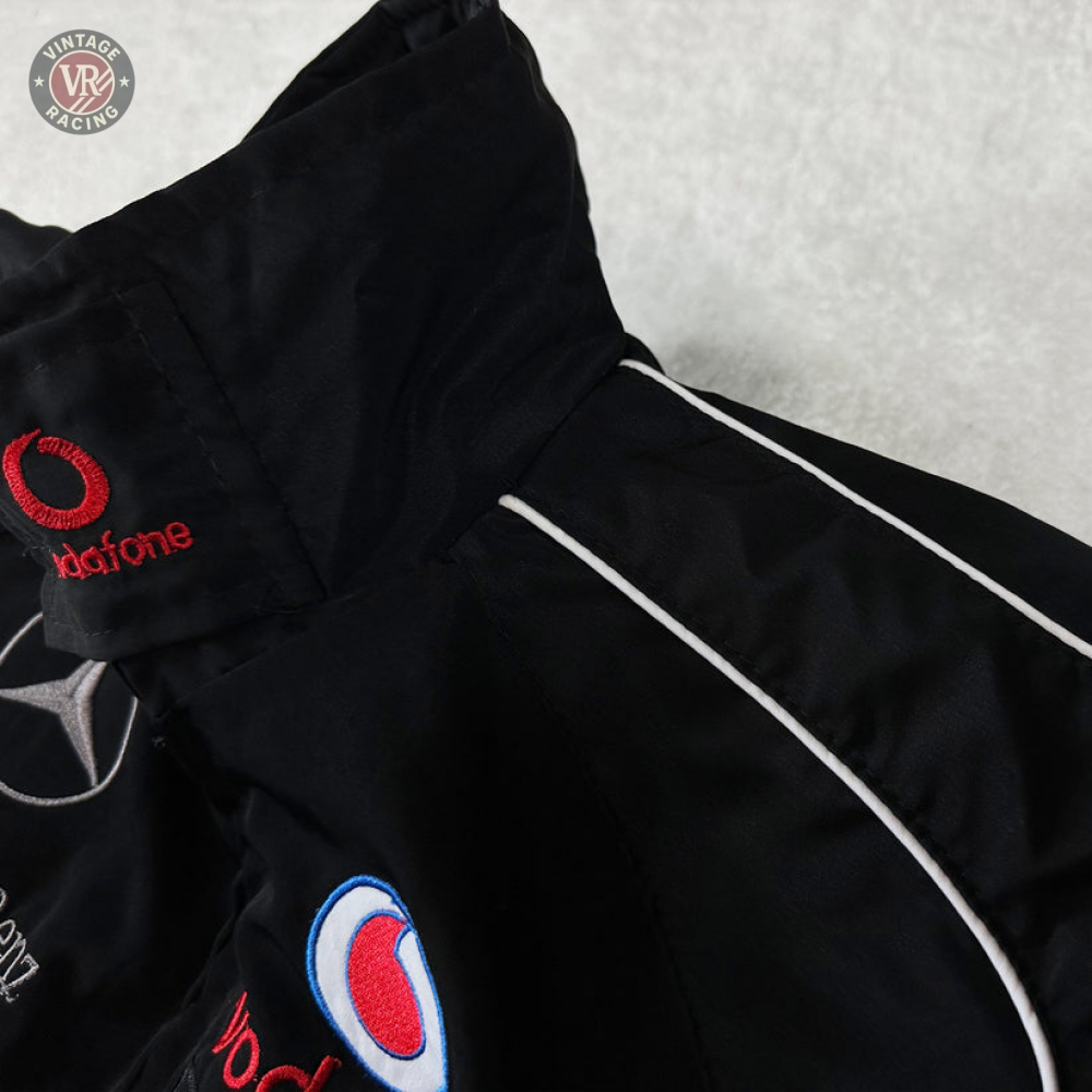 Close-up of the Mercedes-Benz Santander Vintage Jacket in black with white piping, featuring embroidered Vodafone, Mercedes star, and blue-red patches—highlighting its classic motorsport heritage on a light gray background.