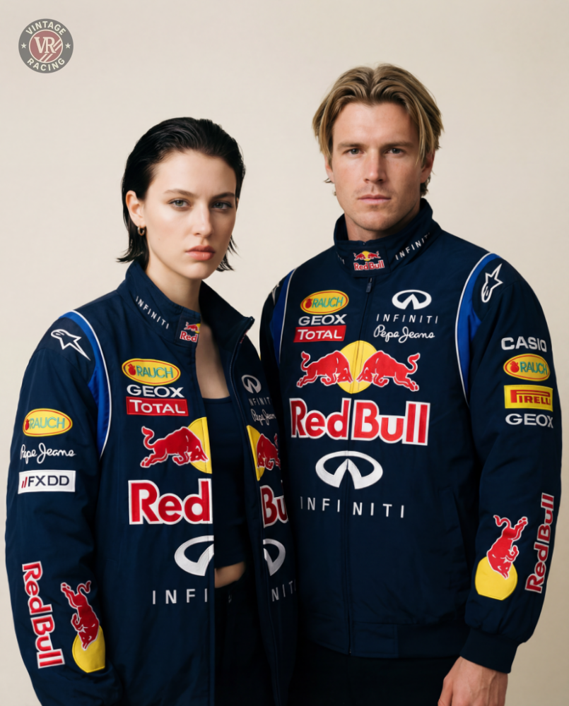 Two people stand side by side against a beige background, looking at the camera in matching Red Bull Racing F1 Jackets, featuring sponsor logos and a navy blue design.