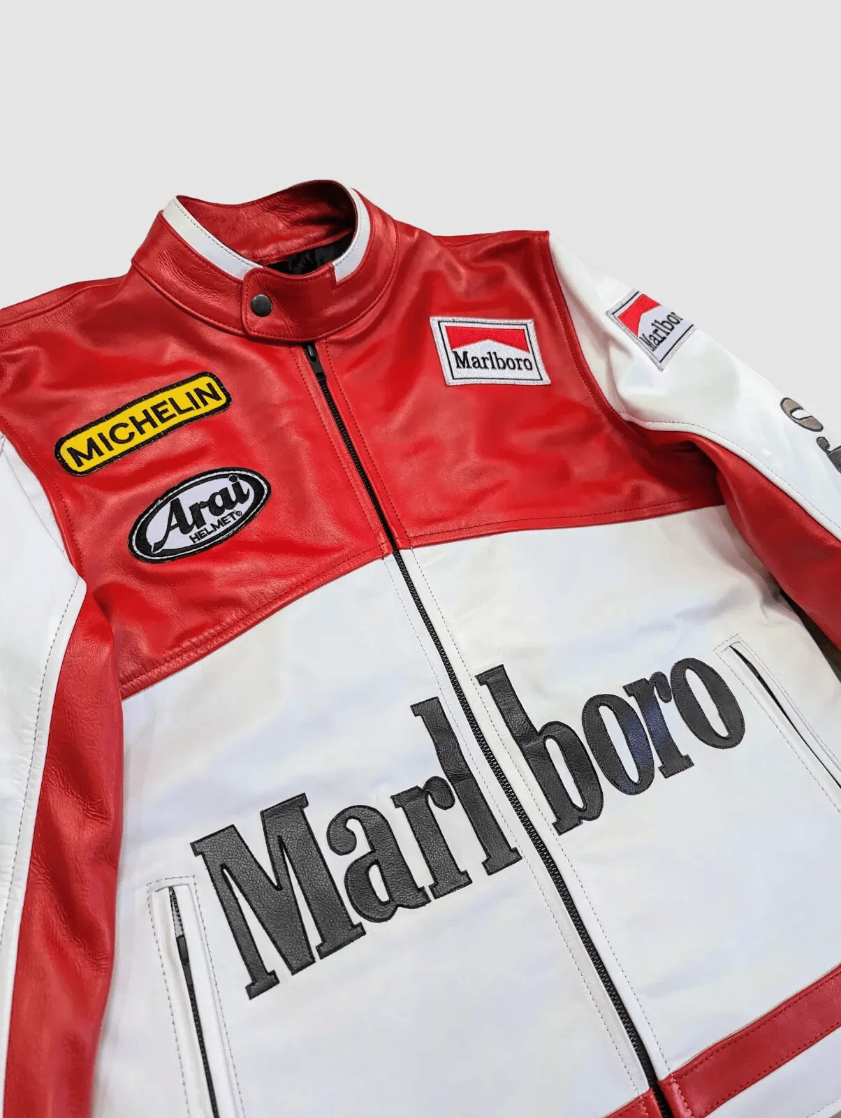The Marlboro Racing Leather Jacket, shown on a light gray background, features bold red and white Marlboro branding with Michelin and Arai patches on the chest and arms for a classic retro motorsport look.