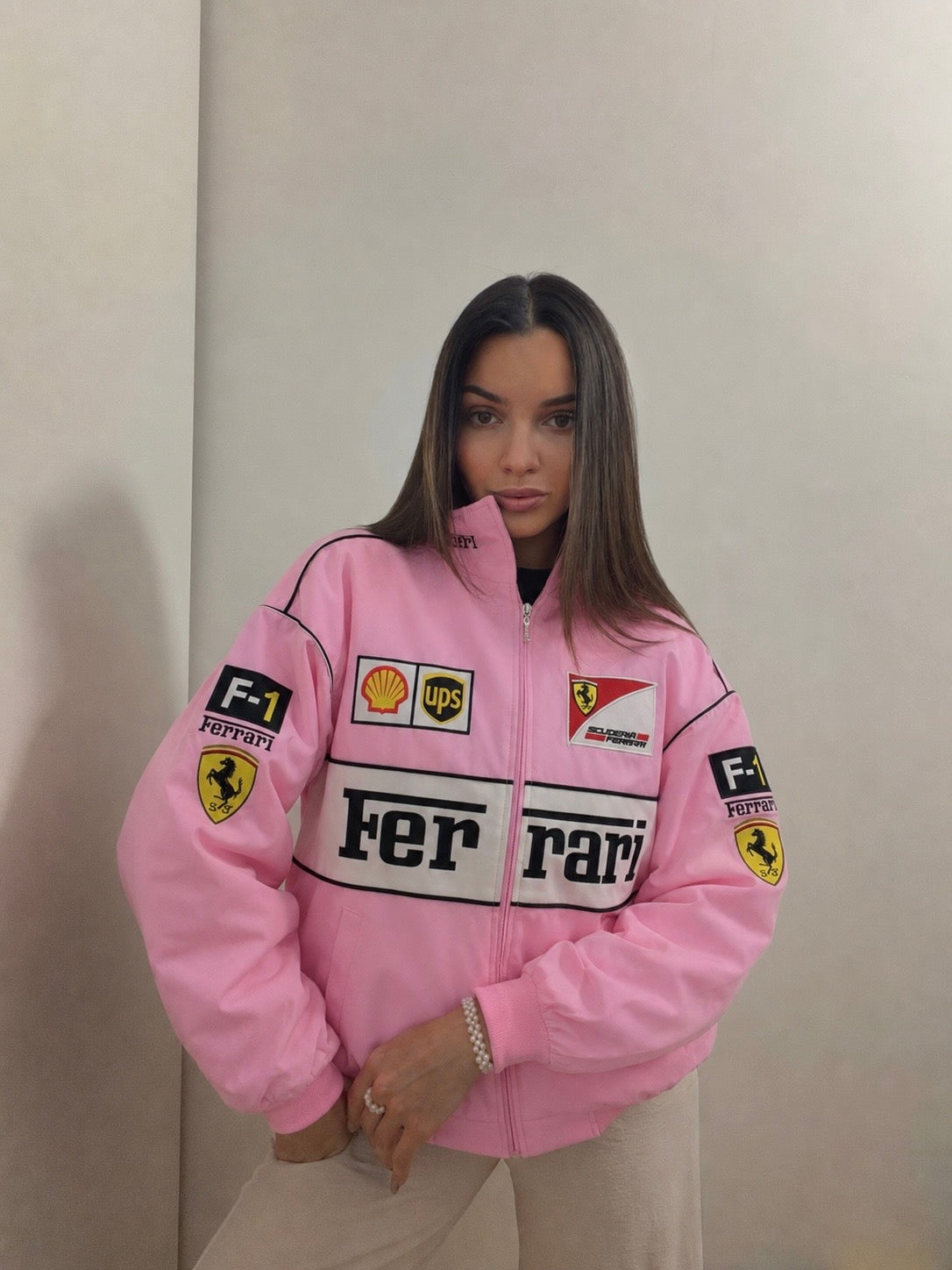 A woman models the Ferrari F1 Vintage Racing Jacket - Pink Edition, featuring various sponsor logos, as she stands indoors against a plain light-colored wall, looking at the camera with her hands in her pockets.