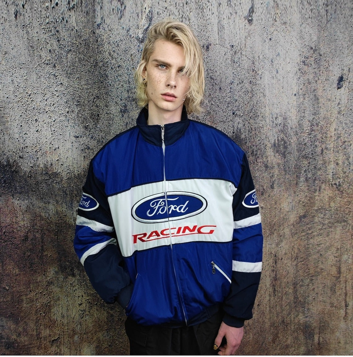 A person with long blonde hair wears the Ford Vintage Jacket - Racing Edition, standing against a textured grey background and looking directly at the camera, evoking classic muscle car heritage.