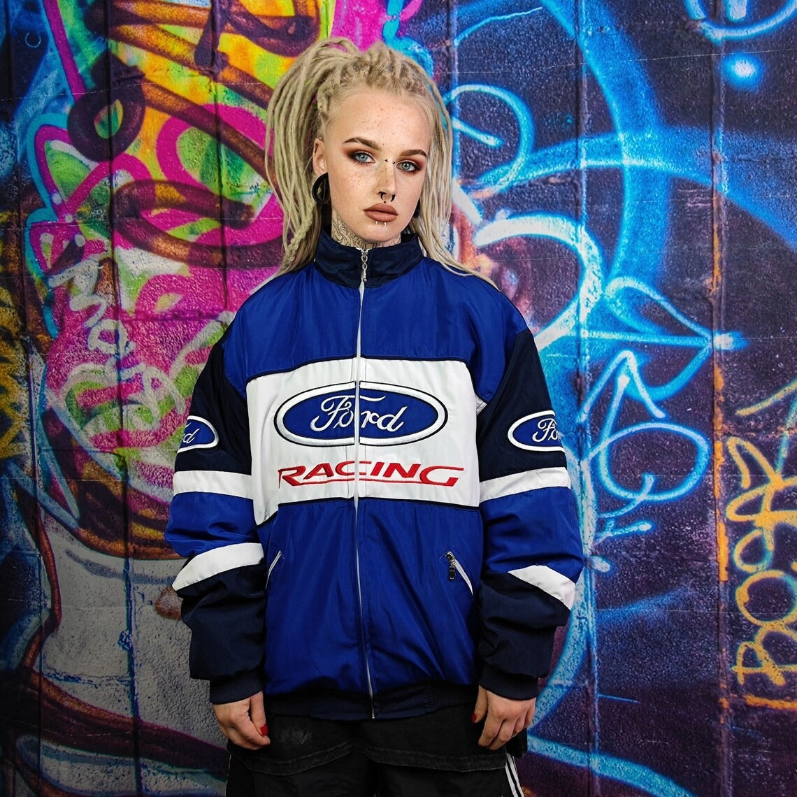 A person with platinum blonde dreadlocks wears the Ford Vintage Jacket - Racing Edition, standing before a vibrant graffiti wall and looking at the camera with a neutral expression, evoking retro motorsport style.