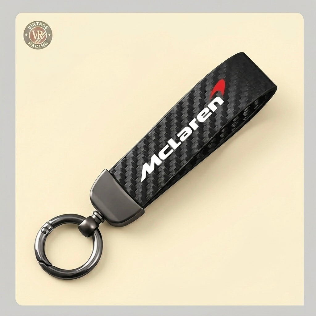 The McLaren Keychain is a black carbon fiber accessory with a metallic ring and features the McLaren logo in white and red on the strap, shown against a light beige background.