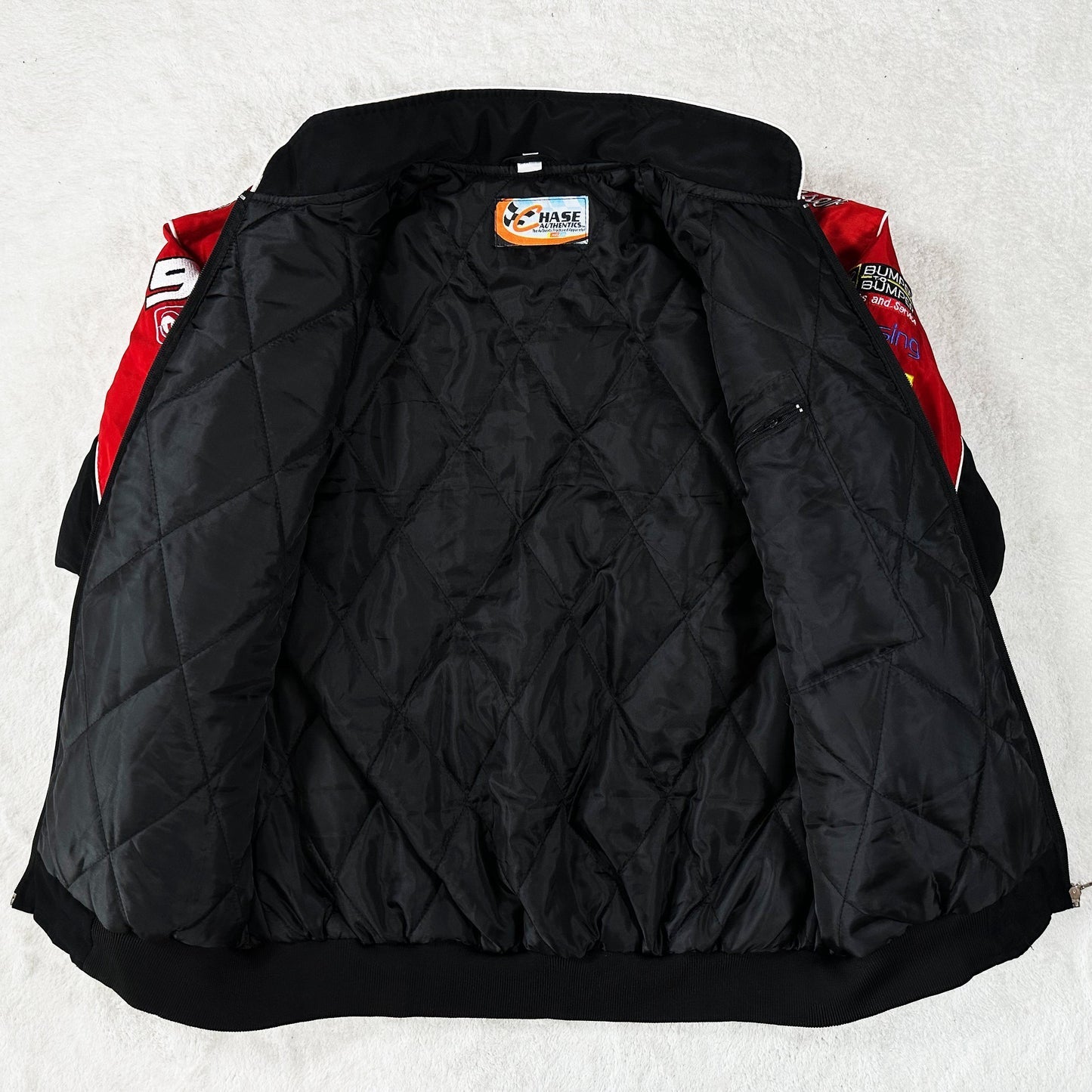 A Budweiser Vintage Racing Jacket - NASCAR Black Edition with a red and black quilted outer layer is spread open on a light surface, displaying its inner lining and a Chase Authentics label at the neck.