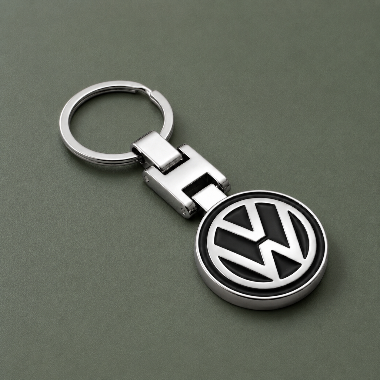 A metallic Volkswagen Keychain featuring a round pendant with the signature VW logo, displayed on a flat greenish-gray surface—perfect for car enthusiasts.