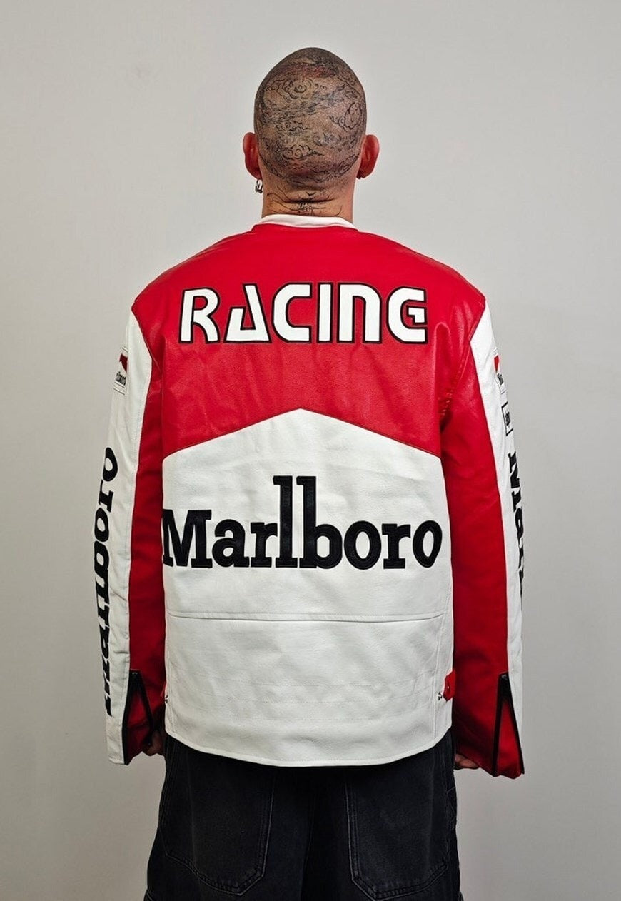 A person with a shaved, tattooed head faces away from the camera, wearing the Marlboro Racing Leather Jacket featuring bold RACING and Marlboro text on the back. The plain light background highlights this striking retro motorsport jacket.