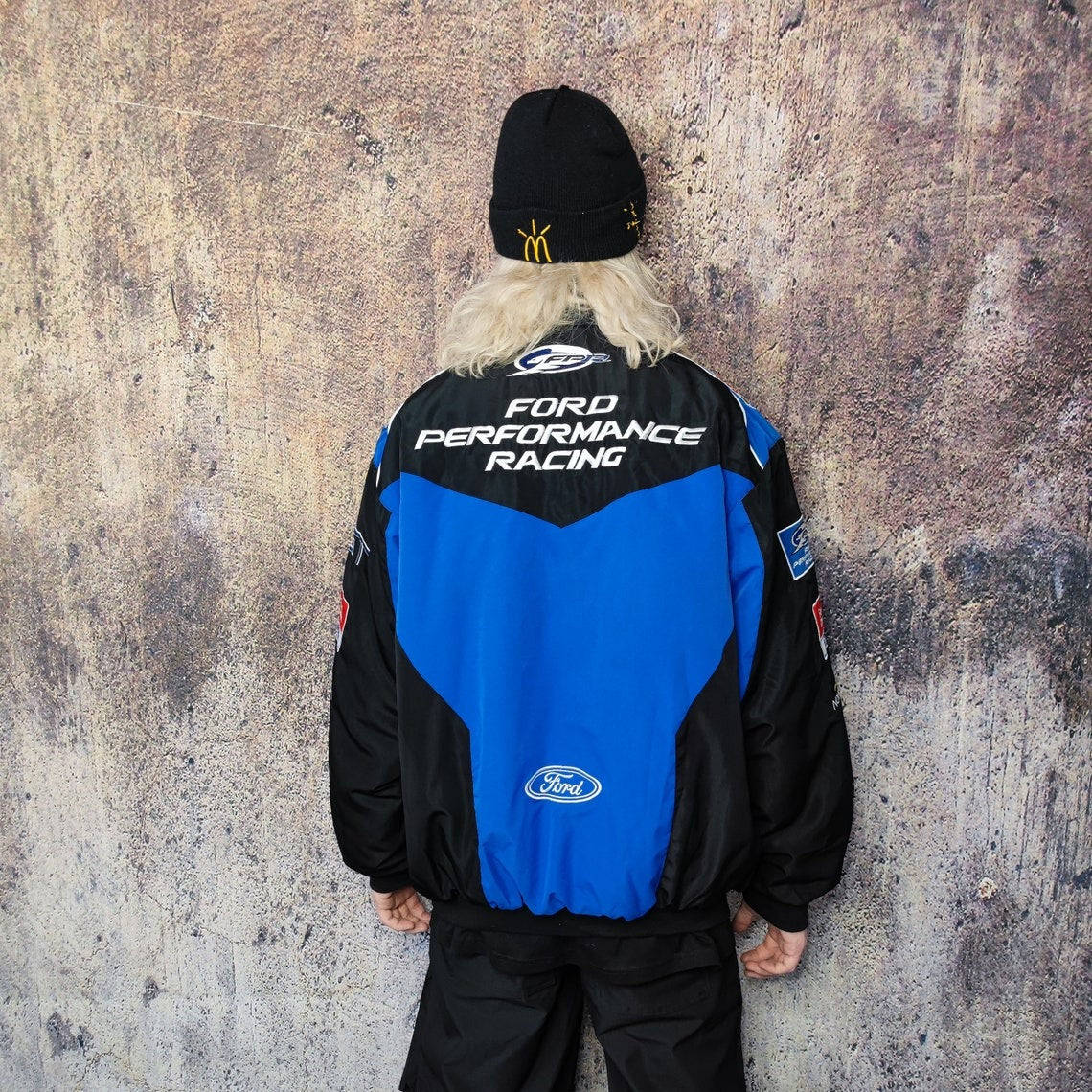 A person with long white hair faces a textured wall, wearing a black beanie and the Ford Vintage Jacket - Performance Edition, which features the iconic Ford logo and celebrates racing heritage in standout streetwear.
