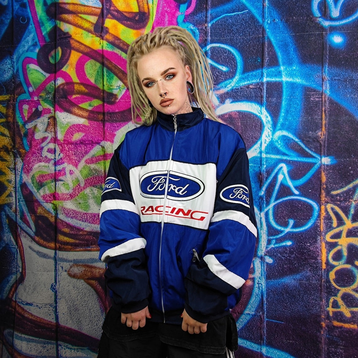 A person with blonde dreadlocks and facial piercings wears a blue Ford Vintage Jacket - Racing Edition, standing before a vibrant graffiti wall, evoking retro motorsport style.
