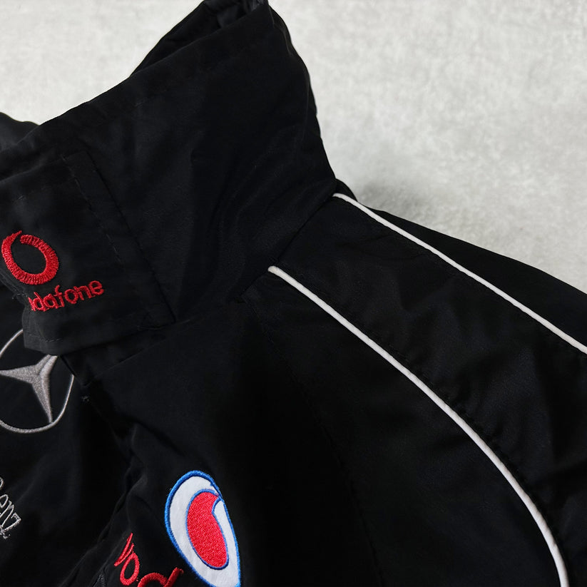 Close-up of the Mercedes-Benz Santander Vintage Jacket in black with white piping, featuring embroidered Vodafone, Mercedes star, and blue-red patches—highlighting its classic motorsport heritage on a light gray background.