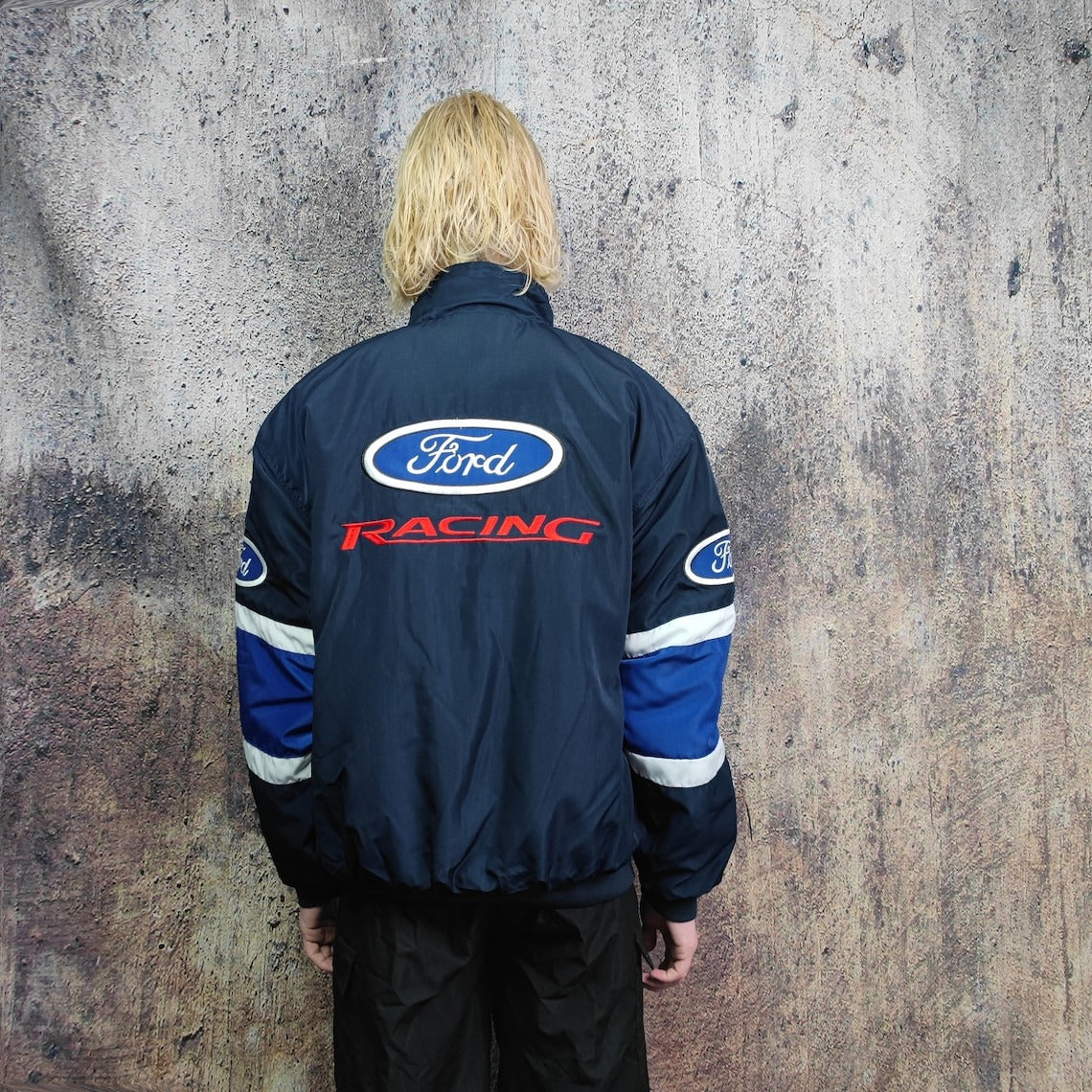 A person with blond hair, seen from behind, wears the Ford Vintage Jacket - Racing Edition with blue and white sleeve stripes, standing against a textured wall—embodying true retro motorsport style.