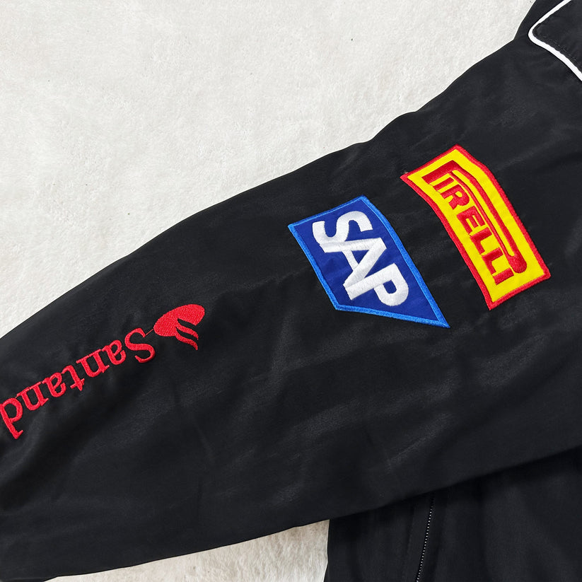 The Mercedes-Benz Santander Vintage Jacket’s black sleeve in stealth racing style showcases embroidered logos: Santander (red), SAP (blue/white), and Pirelli (yellow/red), all set against a light-colored background.
