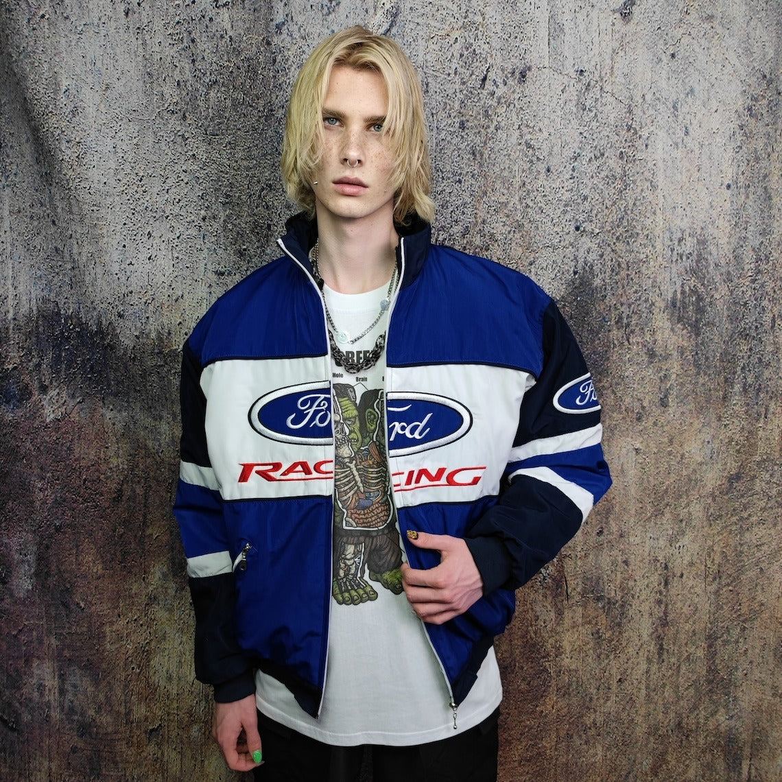 A young person with shoulder-length blonde hair wears the Ford Vintage Jacket - Racing Edition over a graphic tee, standing against a textured wall—capturing the retro motorsport spirit.