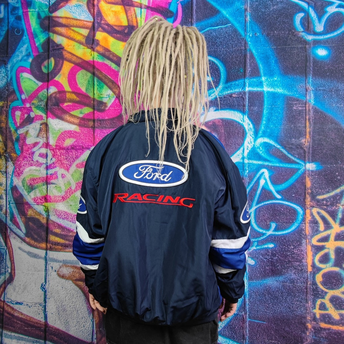 A person with long blonde dreadlocks in a Ford Vintage Jacket - Racing Edition faces a vibrant graffiti wall covered in swirling blue, purple, and pink patterns.