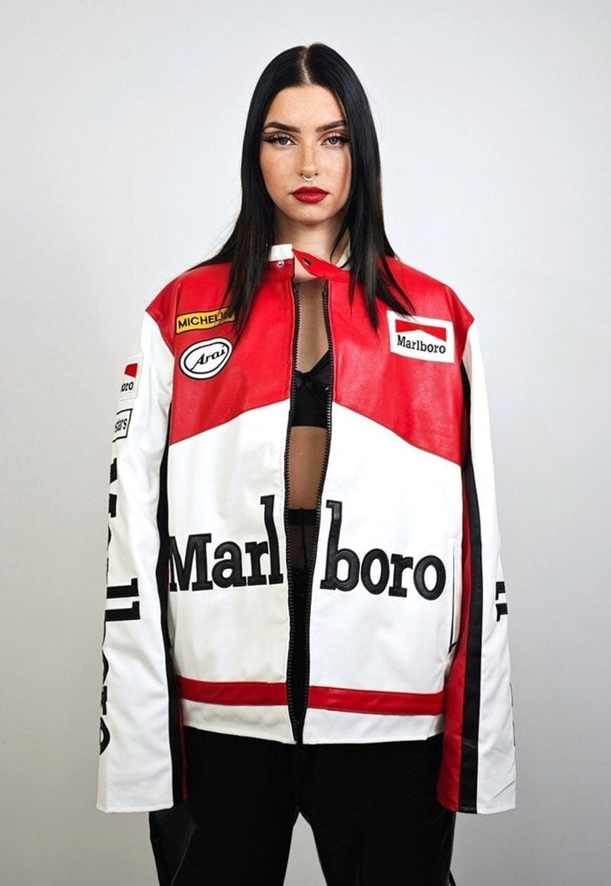 A woman with straight black hair and red lipstick wears the Marlboro Racing Leather Jacket—a bold red, white, and black piece—over a black top, channeling 90s F1 vibes against a plain light background.