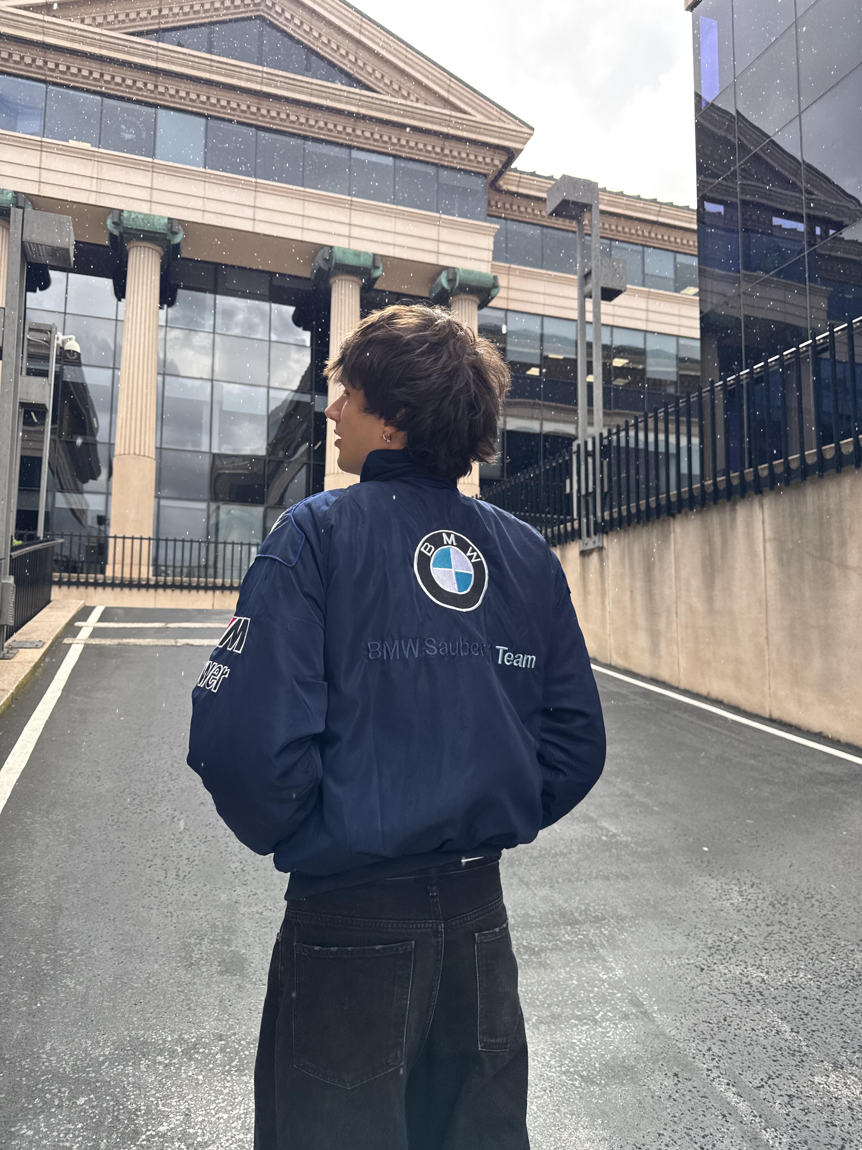 A person with dark hair stands on a paved street facing away, wearing a navy BMW F1 Vintage Racing Jacket - Classic Edition. Modern buildings with large windows and columns rise in the background.
