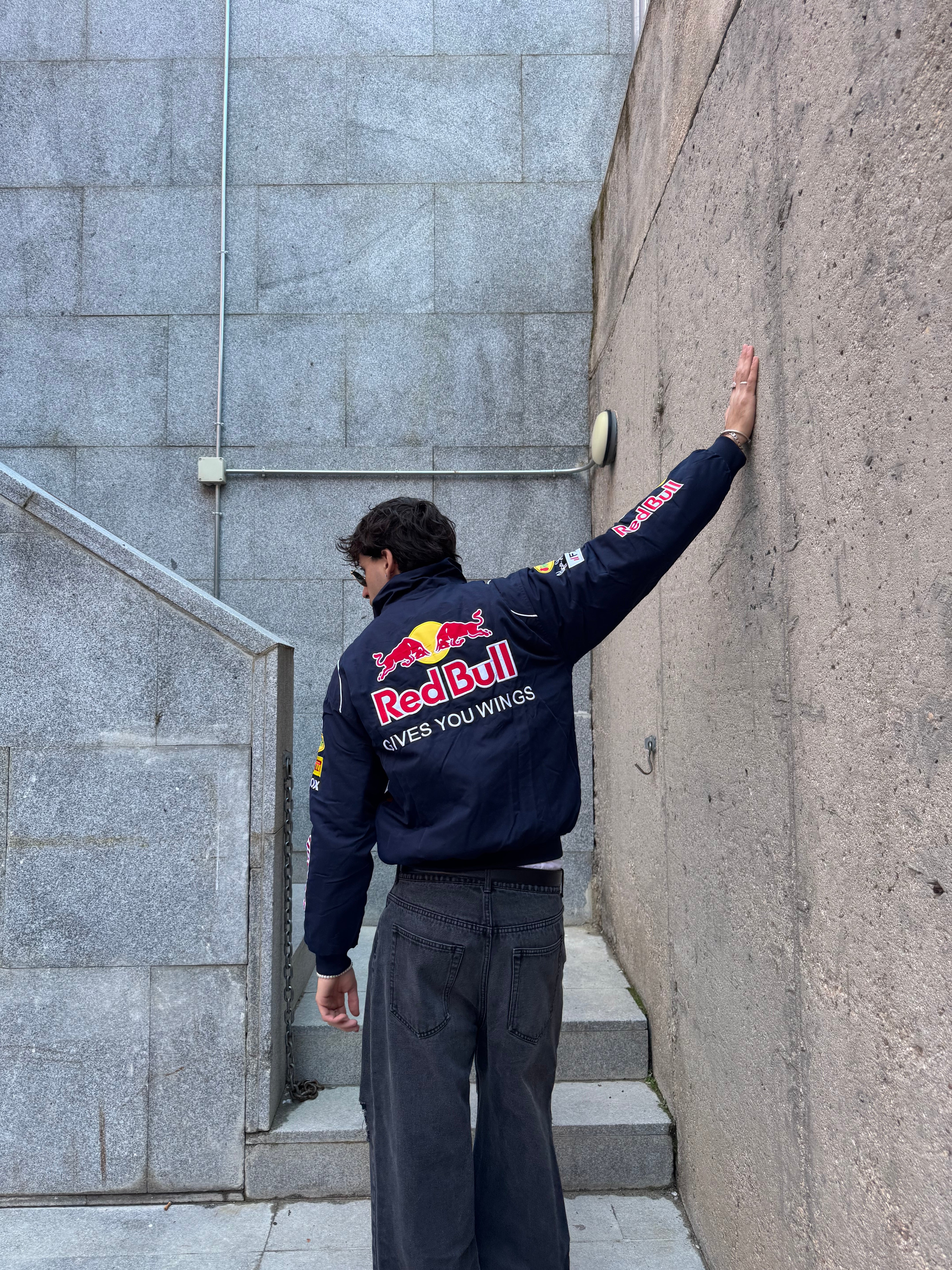 Wearing a Red Bull Racing F1 Jacket, a person stands with one arm extended, touching a concrete wall and facing away from the camera amid stone steps and grey walls in an urban setting.