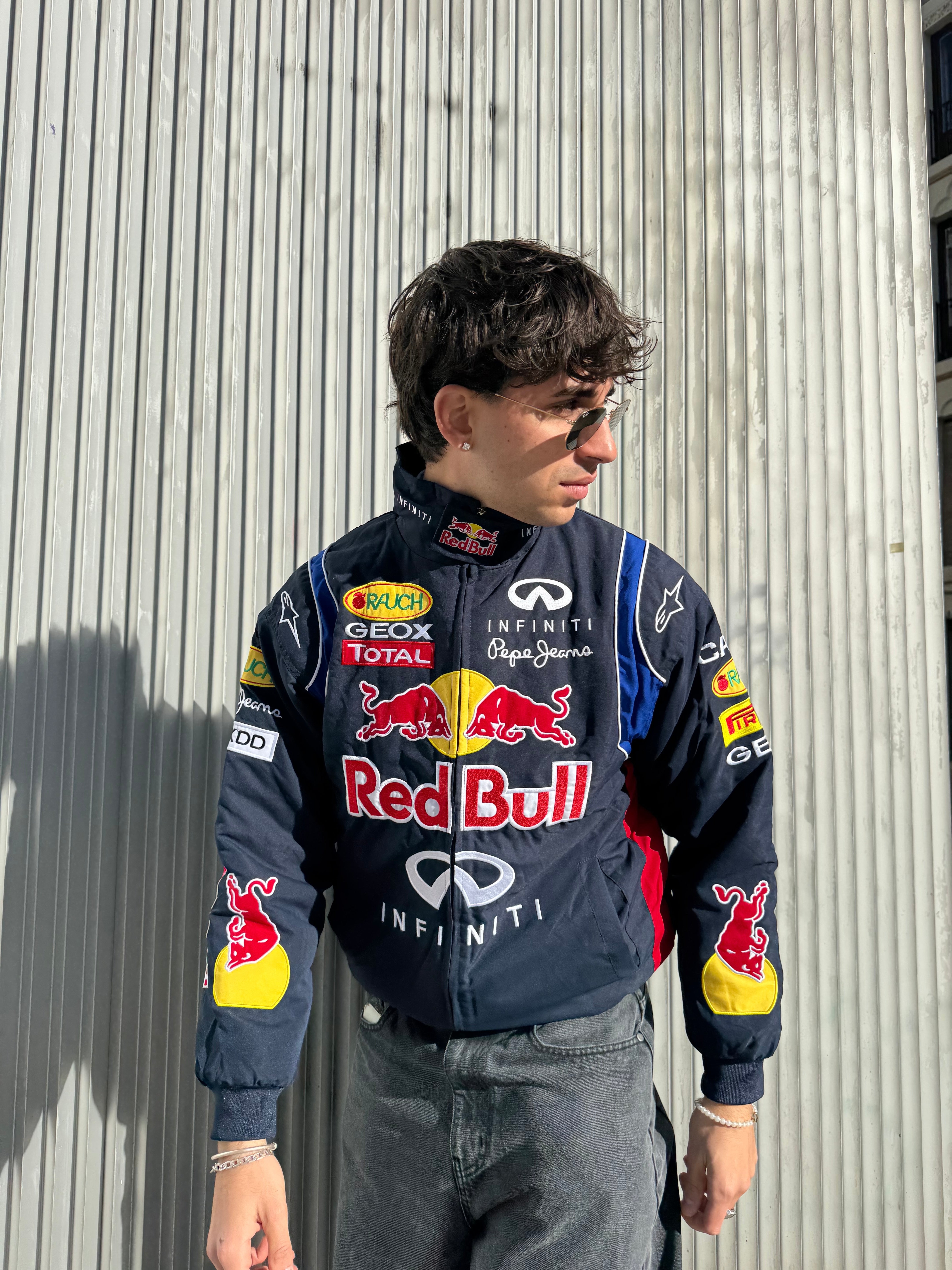 A man in a Red Bull Racing F1 Jacket with sponsor logos and sunglasses stands before a corrugated metal wall, looking to his left.