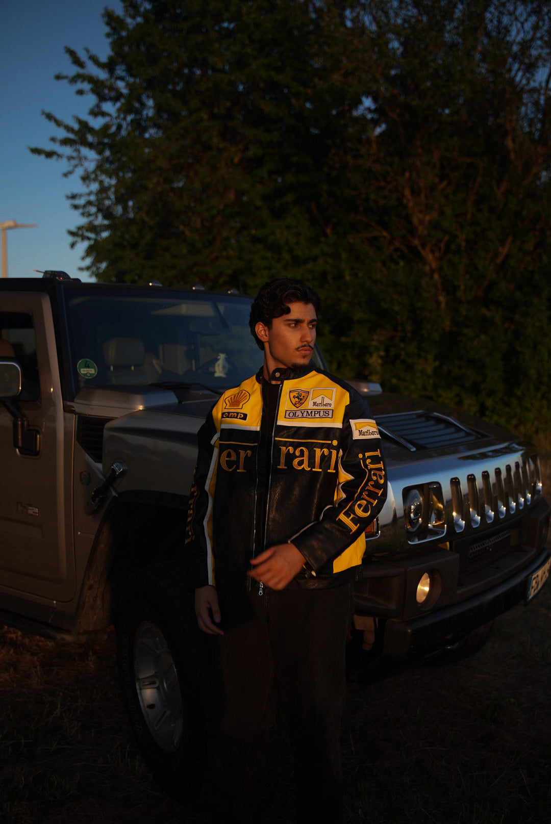 Wearing the Ferrari F1 Vintage Racing Jacket - Black & Yellow Edition, a person stands in front of a gray Hummer SUV outdoors, with trees and sunlight in the background, showcasing a classic retro motorsport style.