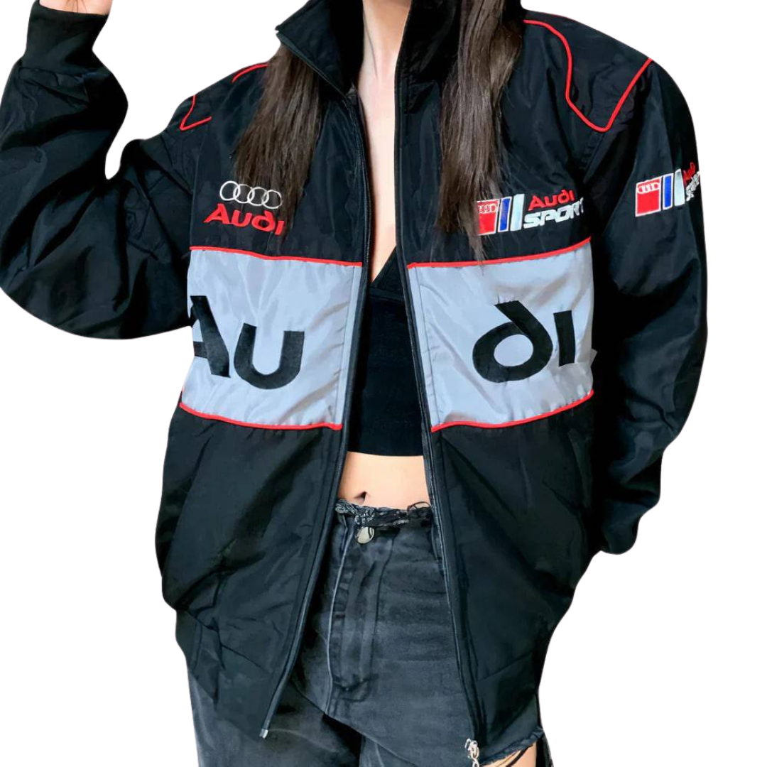 A person wears an Audi Vintage Racing Jacket featuring black fabric with red, white, and gray accents over a black crop top and distressed jeans, standing against a plain white background.