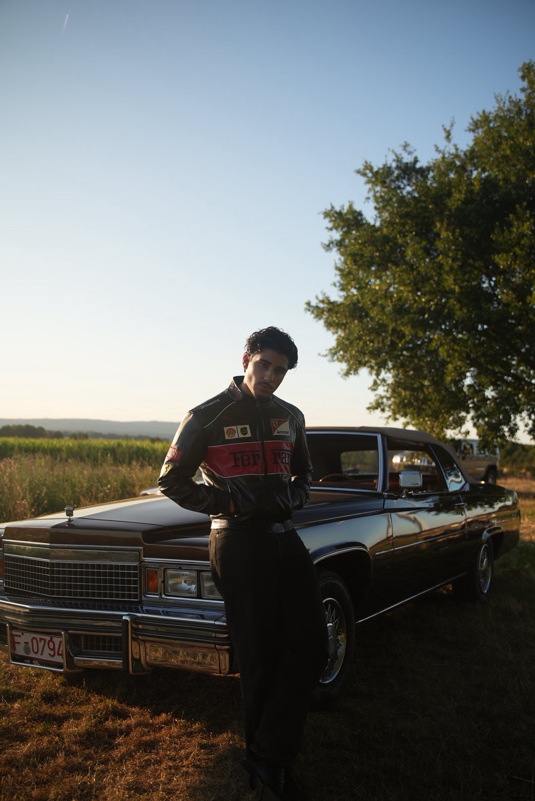 Wearing a Ferrari F1 Vintage Racing Leather Jacket - Red Edition, a person leans against a classic black car parked on grass, framed by a tree and an open field beneath a clear sunset sky.