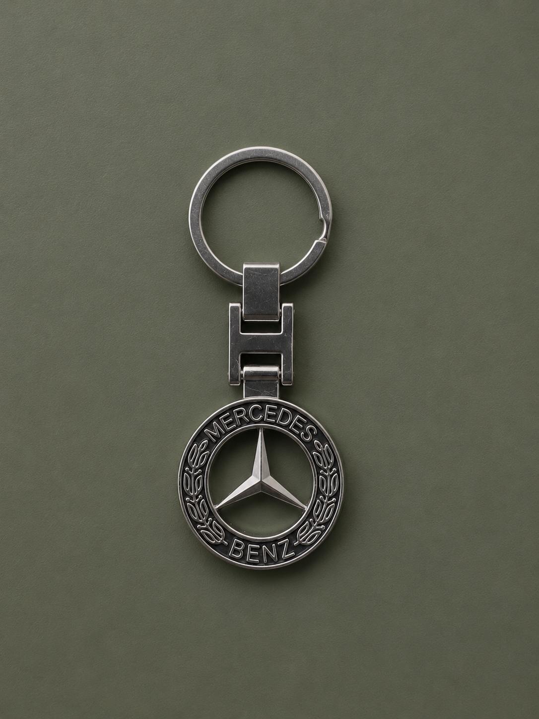 The Mercedes-Benz Keychain showcases the iconic three-pointed star logo with a laurel wreath detail on a plain olive green background.