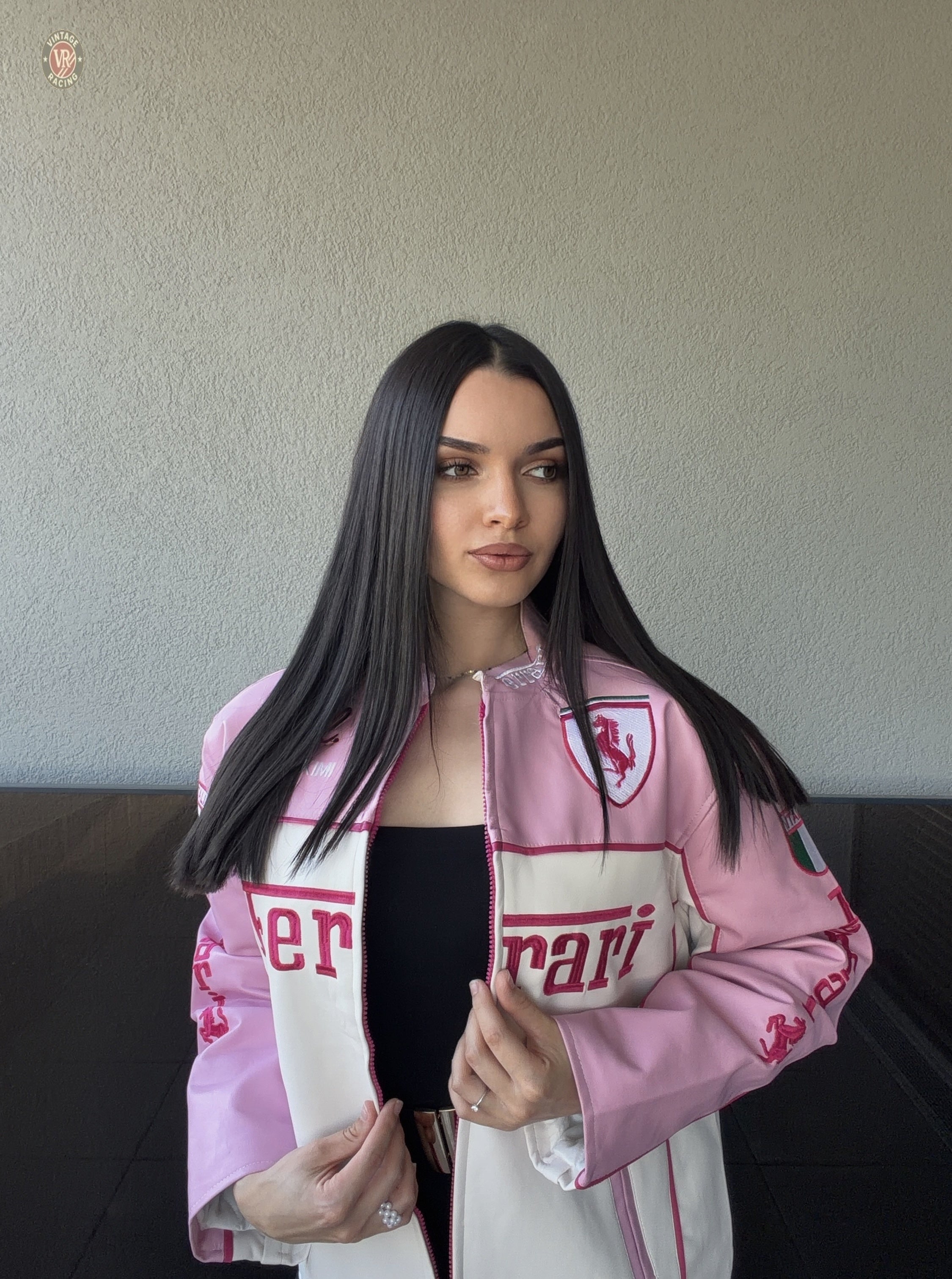 A woman with long straight dark hair, wearing a black top and the Ferrari F1 Vintage Racing Leather Jacket - Pink & White Edition, stands against a plain light wall, gazing slightly to the side with a neutral expression.