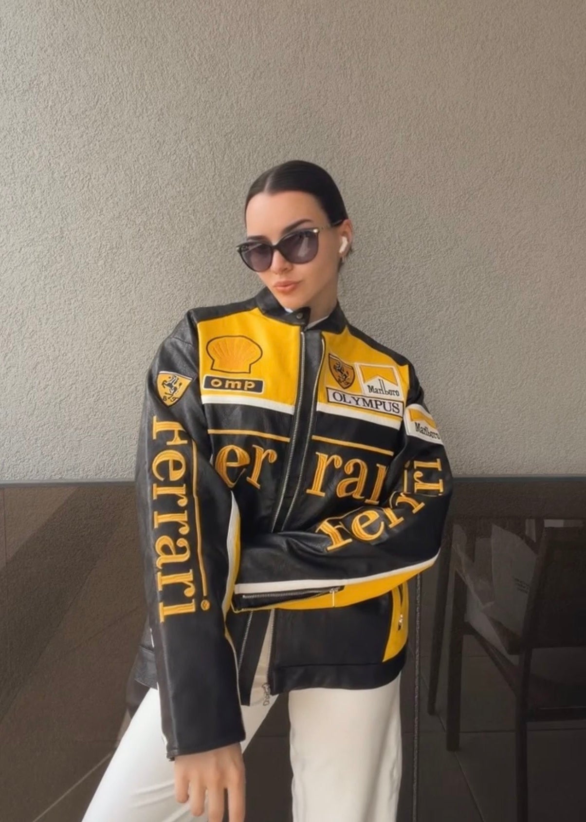 A woman in sunglasses and wireless earbuds poses by a plain wall, styling white pants with the Ferrari F1 Vintage Racing Jacket - Black & Yellow Edition for bold motorsport streetwear vibes.
