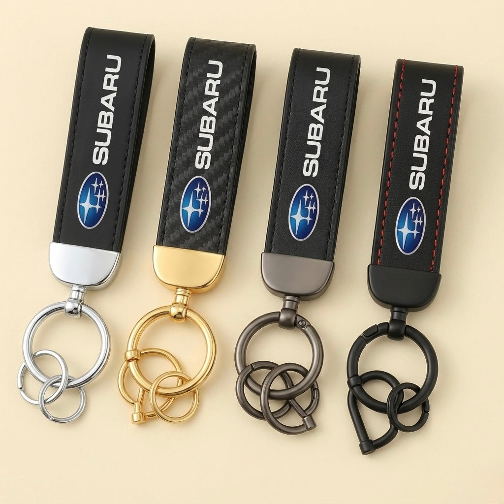 Four Subaru Keychains in silver, gold, gunmetal, and black are displayed side by side. Each features the Subaru logo and a uniquely colored metal keyring, making them stylish accessories against a beige background.