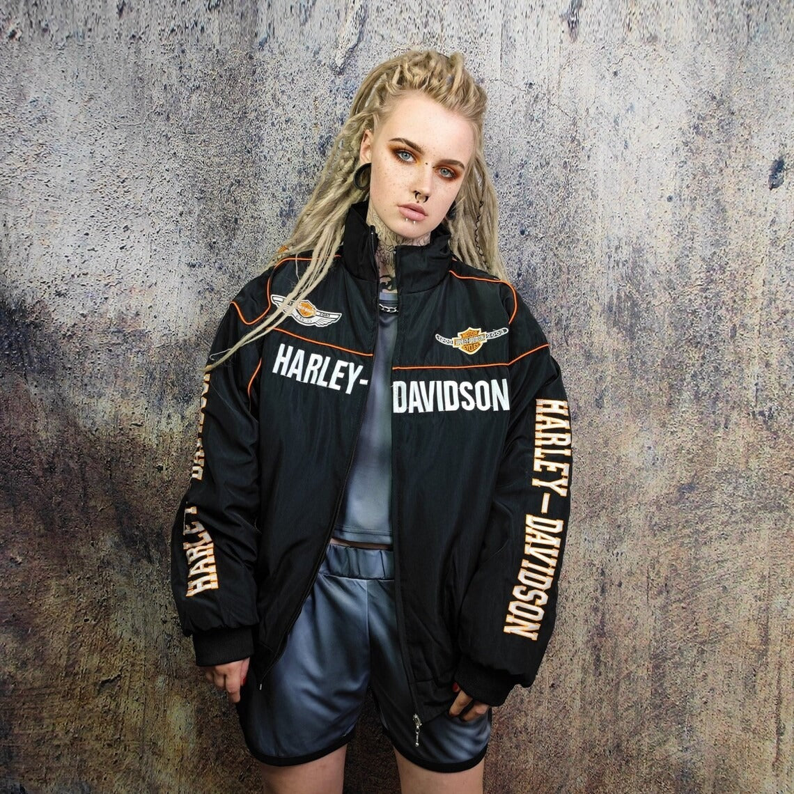 Wearing a Vintage Harley Davidson Jacket over a shiny gray outfit, a person with long blonde dreadlocks stands against a speckled wall, looking directly at the camera with a neutral expression.