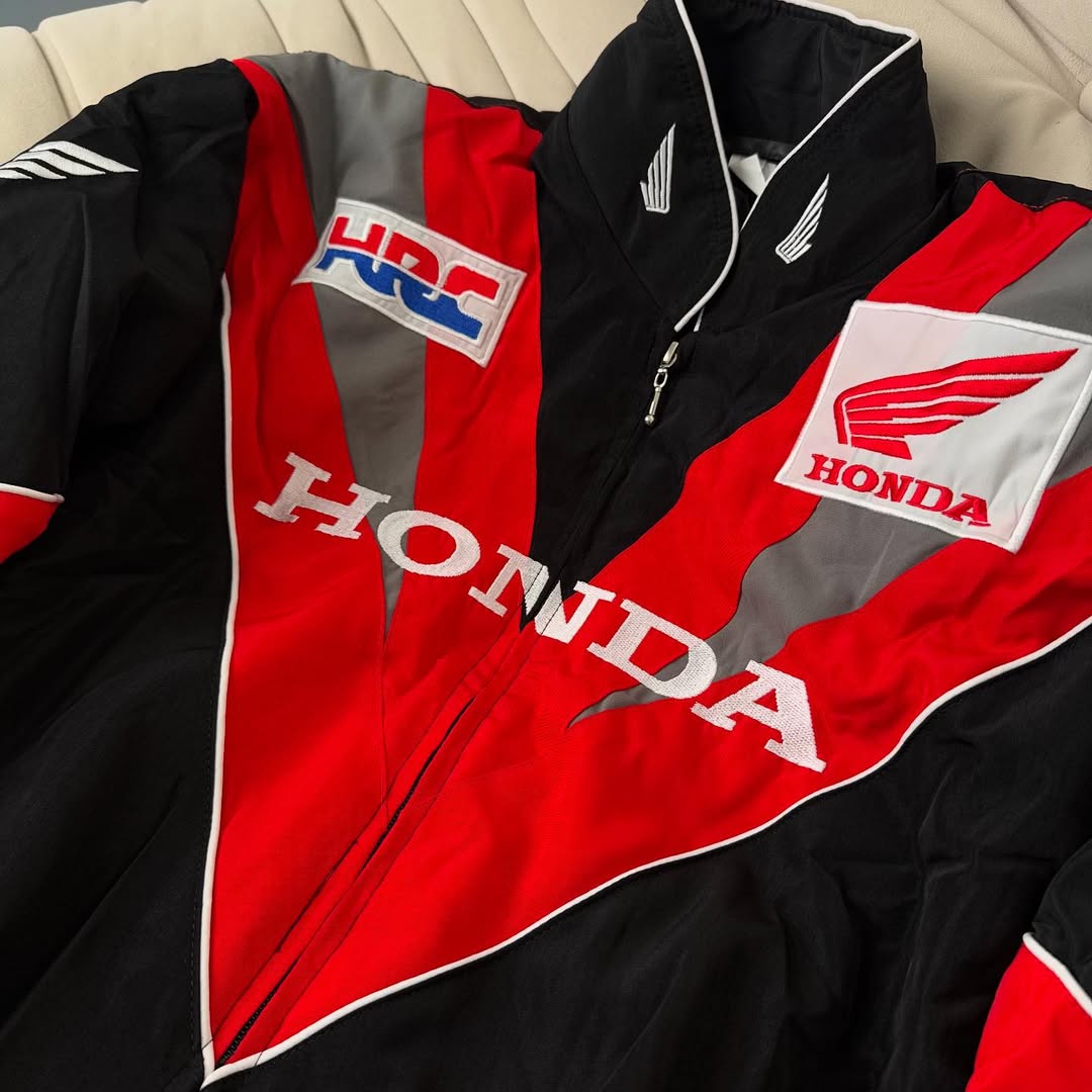 A close-up of the Honda Vintage Racing Jacket in black, red, and white with bold HONDA lettering, wing logos, and an HRC patch is displayed on a beige surface.