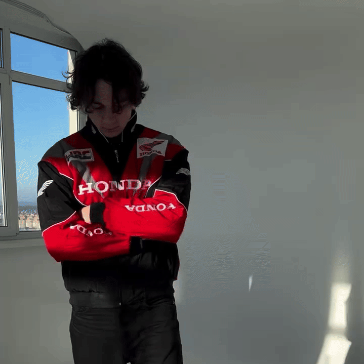 Indoors near a sunlit window, a person with wavy dark hair stands with arms crossed, wearing the Honda Vintage Racing Jacket in red and black.