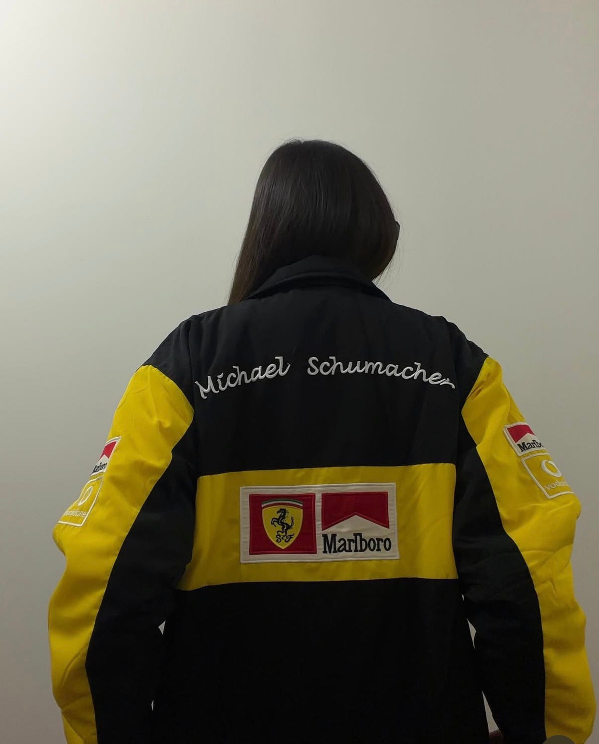 A person wearing a black and yellow Vintage Ferrari Michael Schumacher Jacket, featuring the name Michael Schumacher, a shield logo, and sponsorship branding on the back.