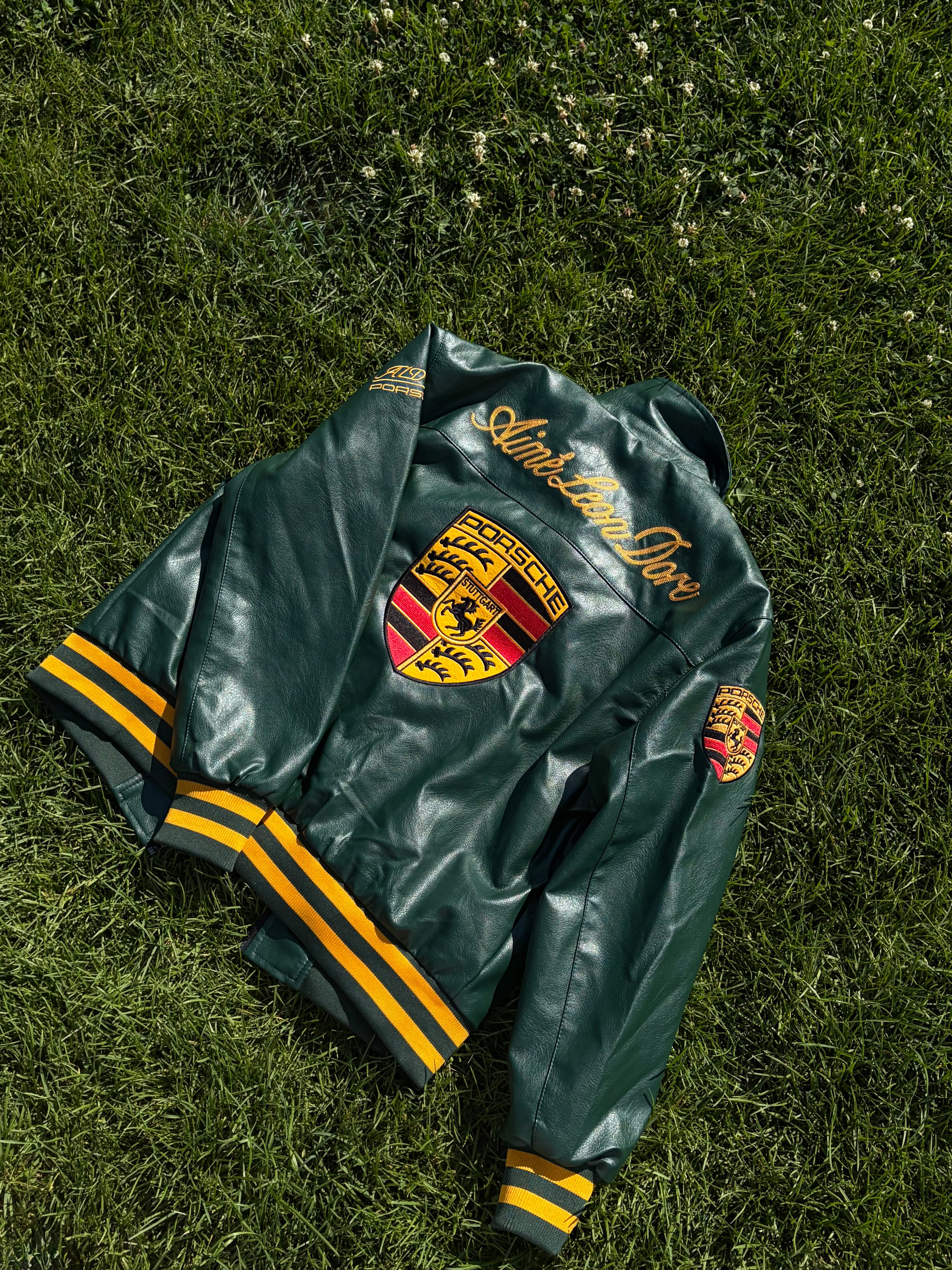 The Porsche Vintage Leather Bomber Jacket - Green Edition, featuring yellow accents and Porsche logos, rests on the grass and channels classic motorsport heritage. “Automobilclub” is emblazoned across the back, with similarly branded patches adorning the sleeves.