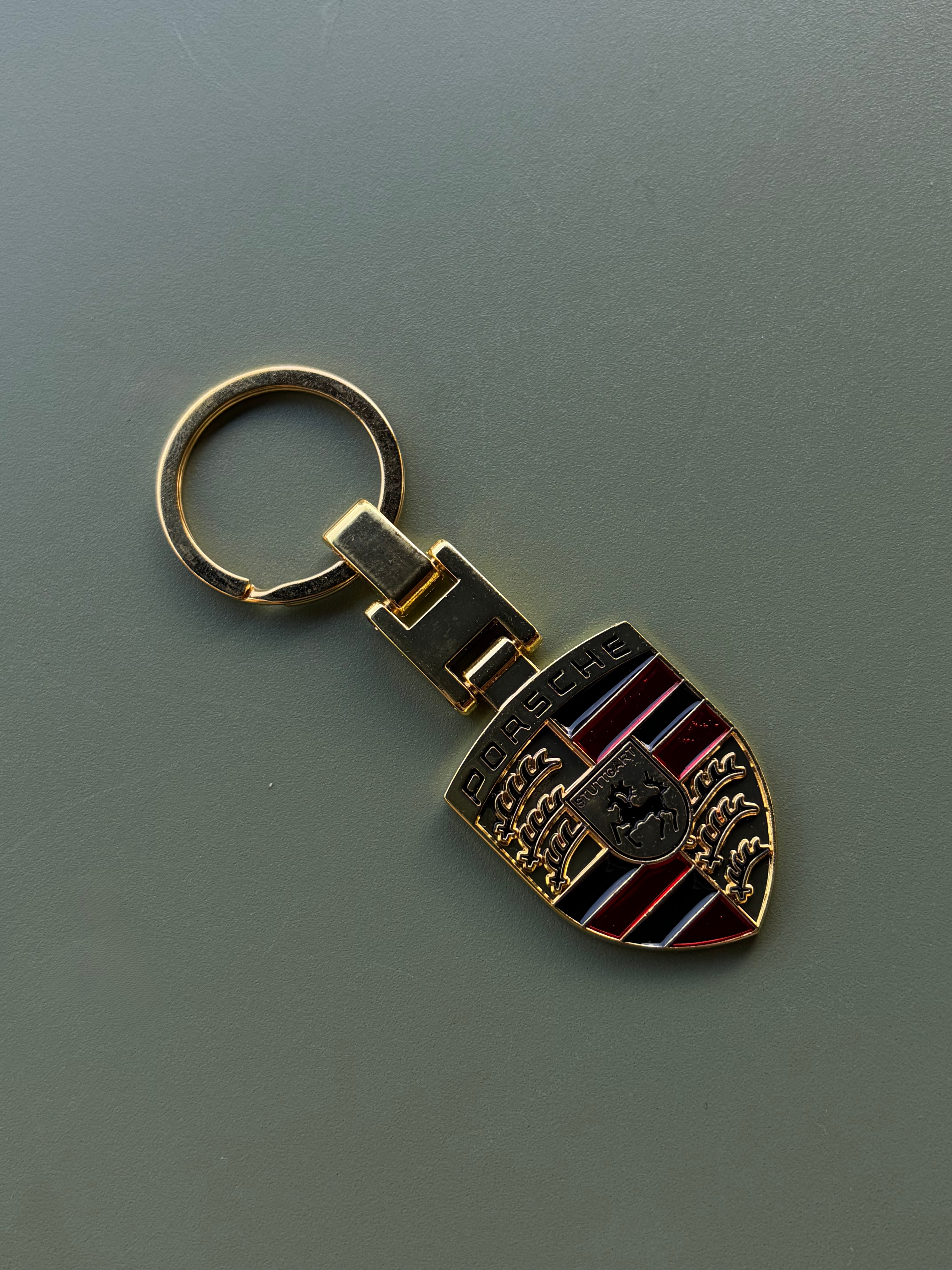 The Porsche Keychain showcases the iconic Porsche crest—with a horse and bold red and black stripes—resting on a flat gray surface, making it the perfect gift for any Porsche enthusiast.