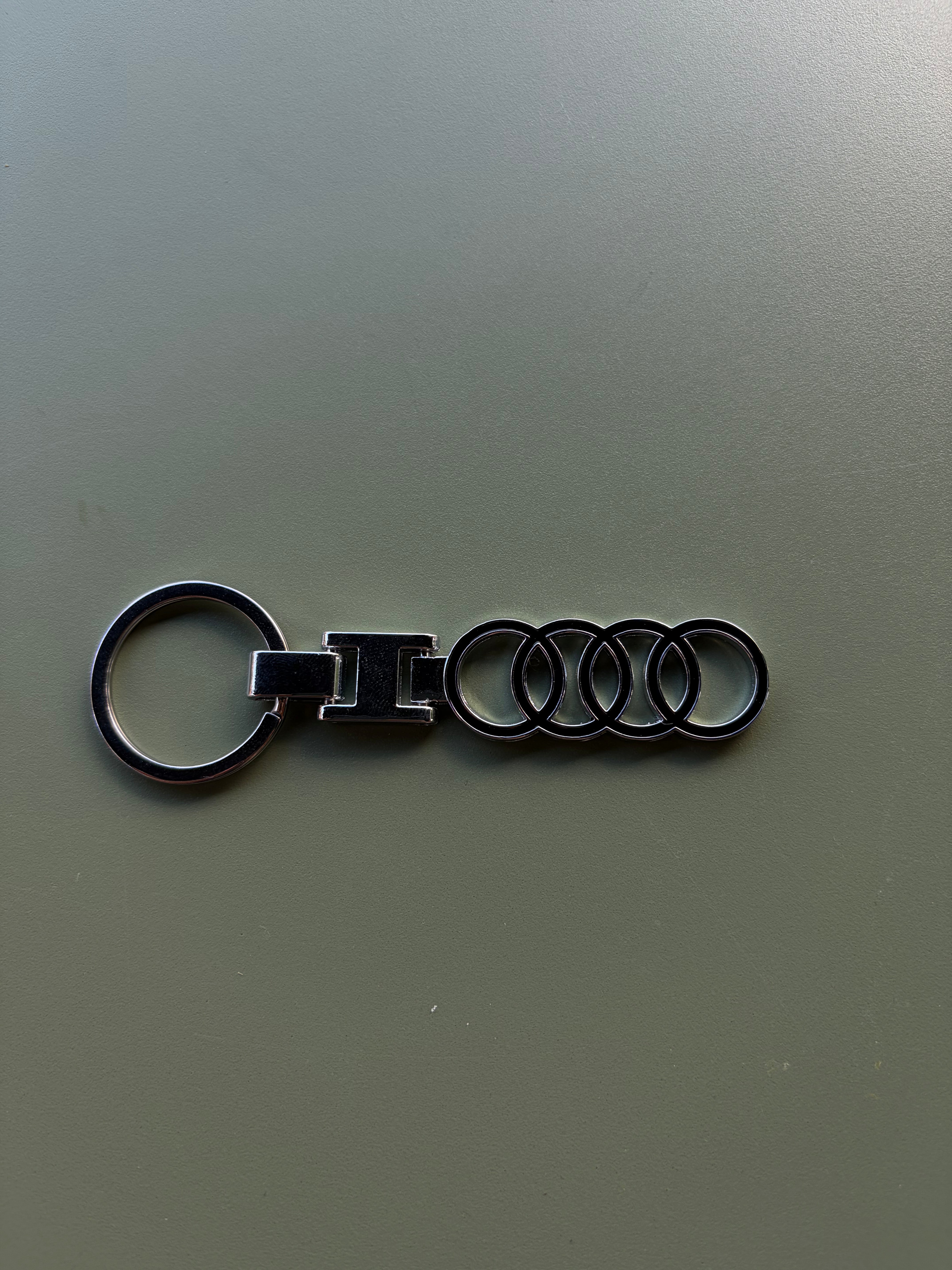 The Audi Keychain, crafted in polished silver and featuring the distinctive four interlinked rings of the iconic Audi logo, rests elegantly on a plain gray surface.