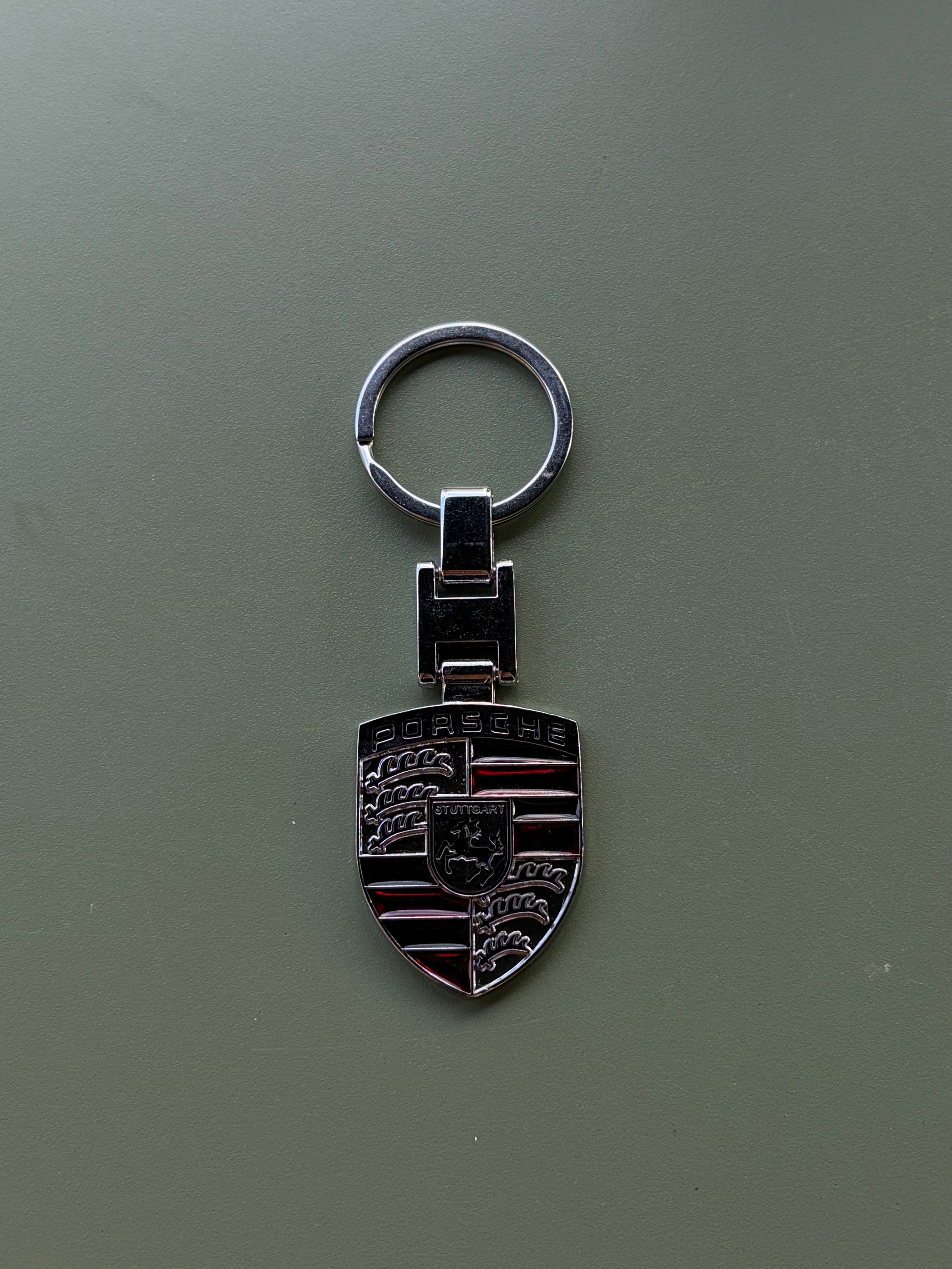 A Porsche Keychain featuring the iconic shield crest with red, black, and gold details rests on a smooth matte green surface. The top of the logo displays the word “Porsche,” making this keychain a perfect gift for fans and collectors alike.