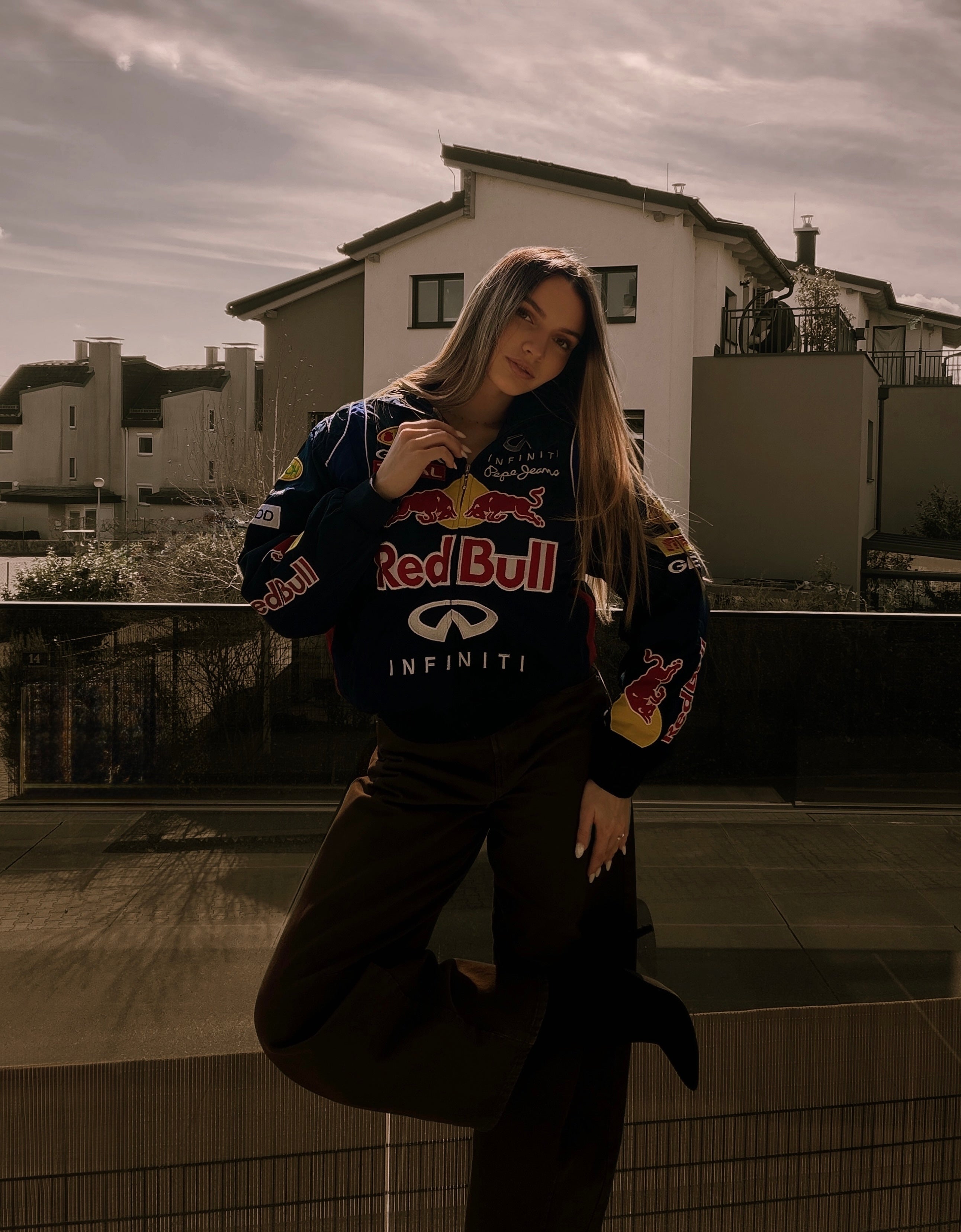 A woman poses outdoors in front of modern houses, wearing the Red Bull Racing F1 Jacket—a standout piece of fan apparel—and dark pants, with one leg slightly lifted and her hand touching her hair. The lighting is soft and warm.