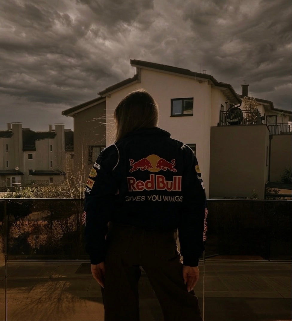 A person wearing the Red Bull Racing F1 Jacket stands on a balcony, facing cloudy skies and modern houses with their back to the camera.