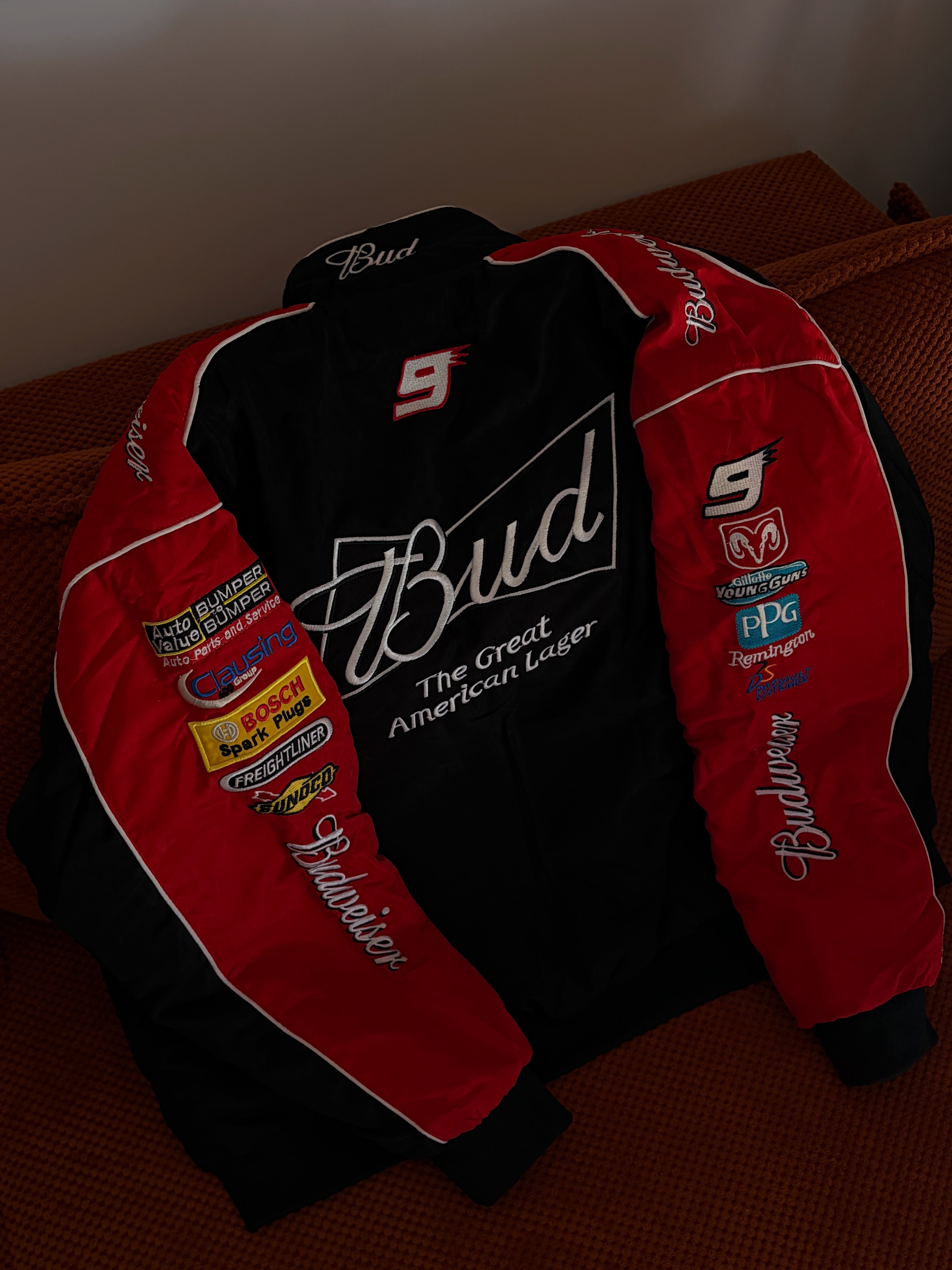 A Budweiser Vintage Racing Jacket - NASCAR Black Edition, in classic black and red with sponsor patches and “Bud The Great American Lager” text, lies spread out on a brown textured surface—an iconic retro piece perfect for any racing enthusiast.