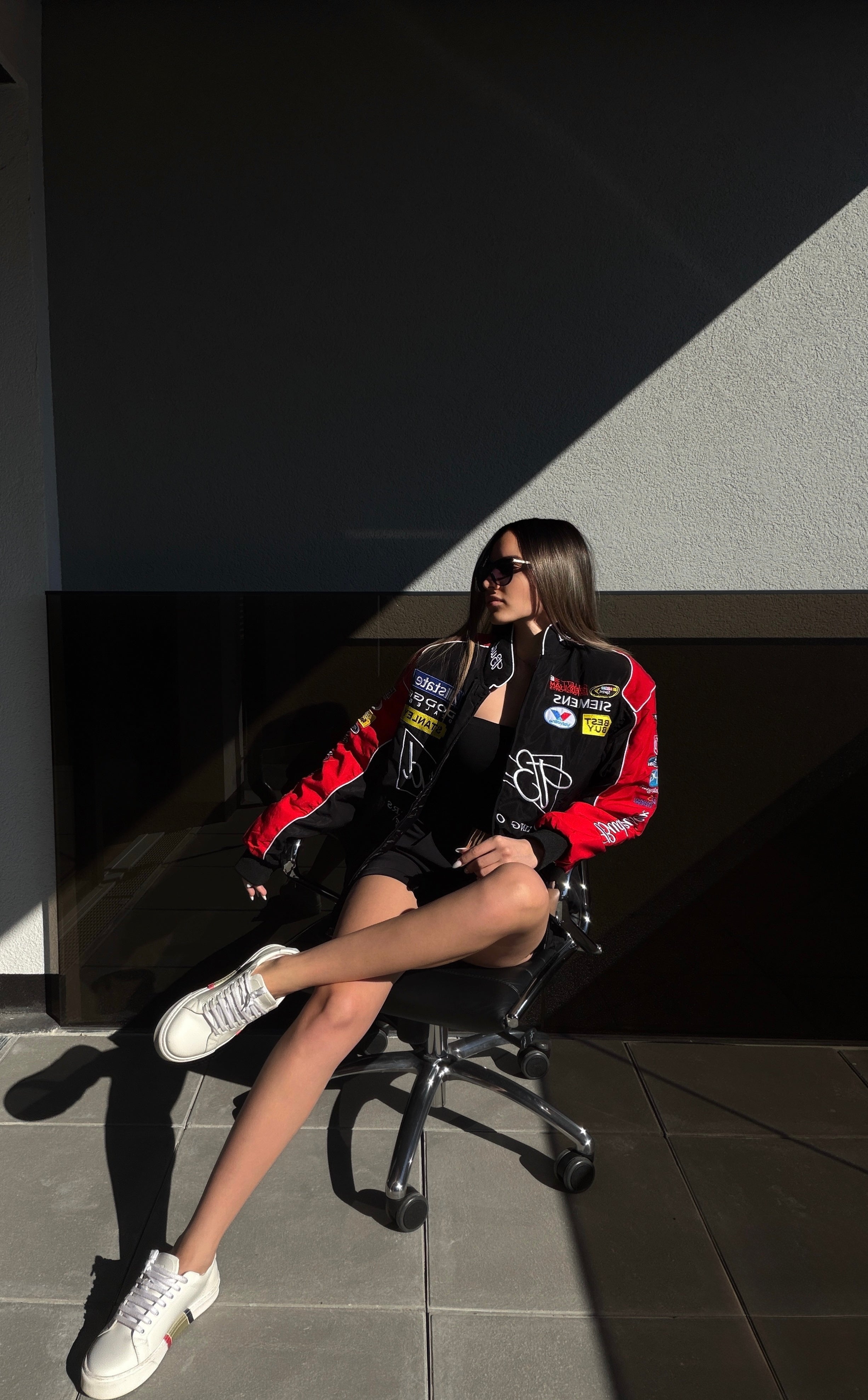 A woman sits in a chair wearing sunglasses, a black skirt, white sneakers, and the Budweiser Vintage Racing Jacket - NASCAR Black Edition with bold red and black patches, as sunlight casts shadows on the wall and floor.
