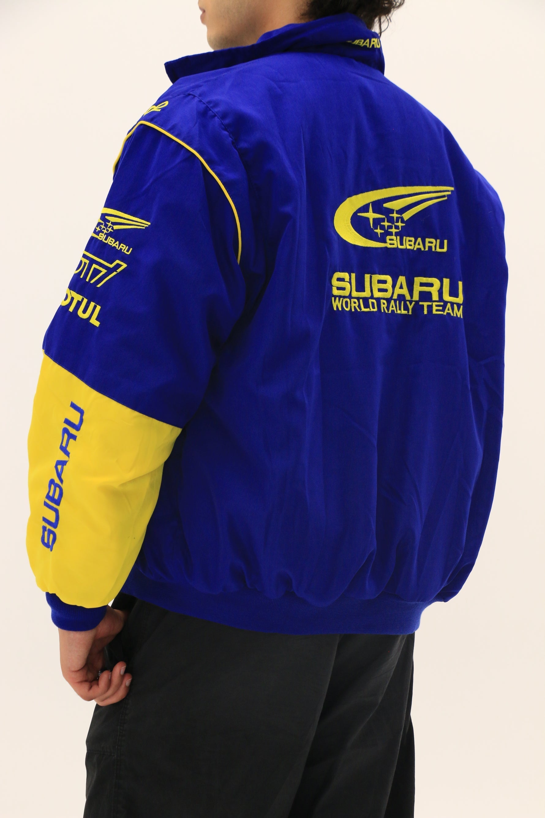 A person wearing the Subaru World Rally Team Vintage Jacket in blue and yellow 555 livery, featuring a large Subaru logo and text on the back and left sleeve, stands against a plain white background.