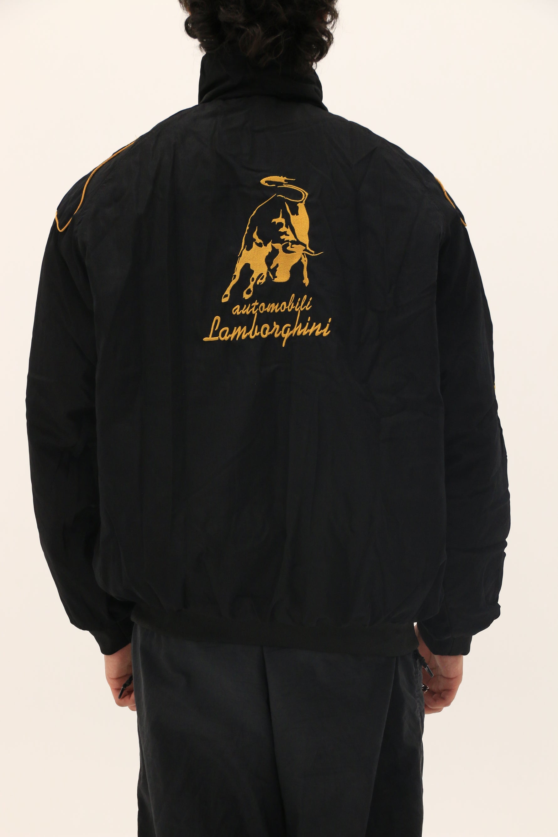 A person faces away wearing the Lamborghini Vintage Racing Jacket, black with gold stitching, a bull, halo, and “automobili Lamborghini” in cursive—ideal for motorsport fans who love retro racing style.