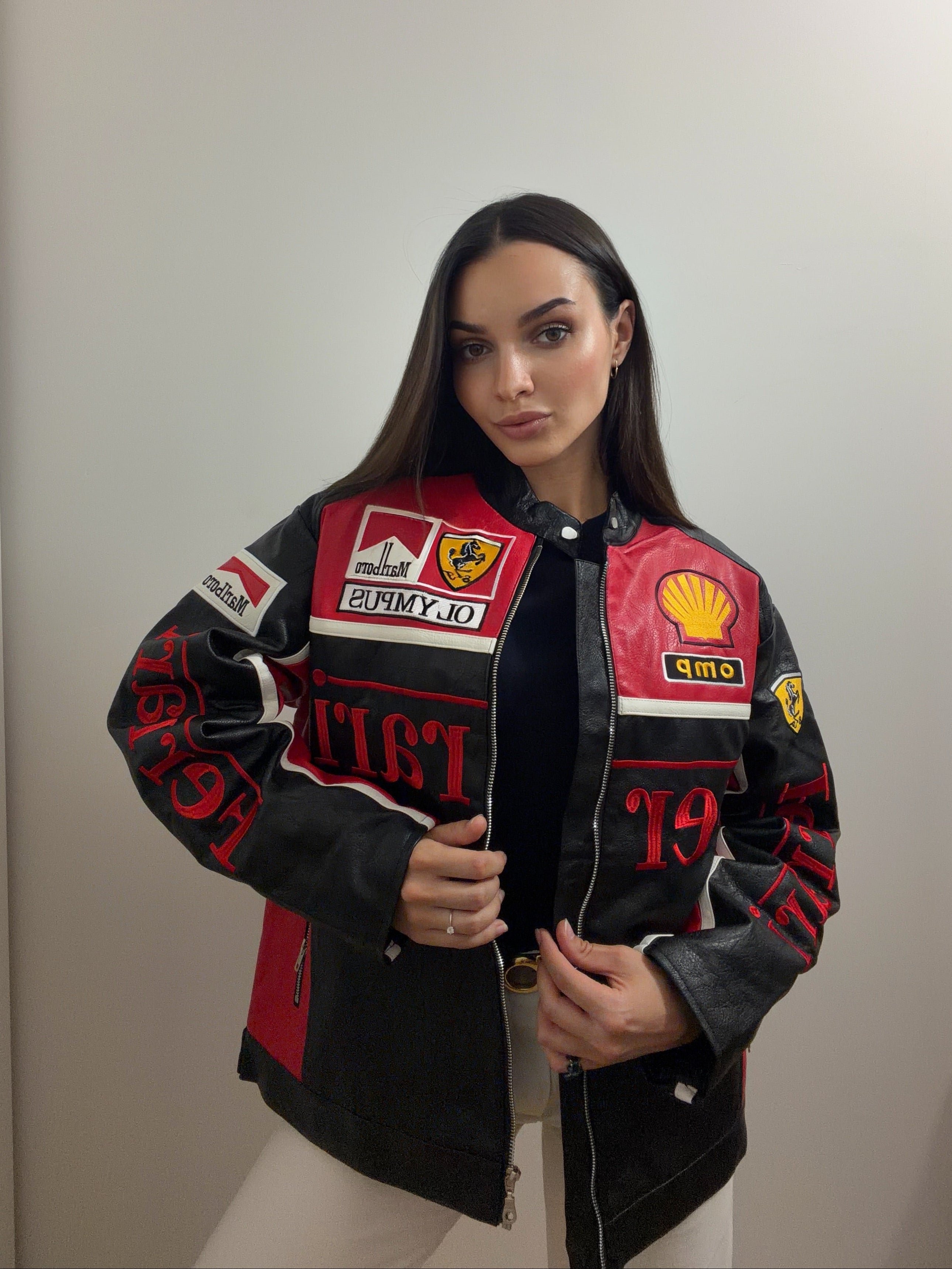 A woman poses against a plain wall wearing the Ferrari Leather Racing Jacket in black and red, adorned with various sponsor patches, paired with white pants, and looks confidently at the camera.