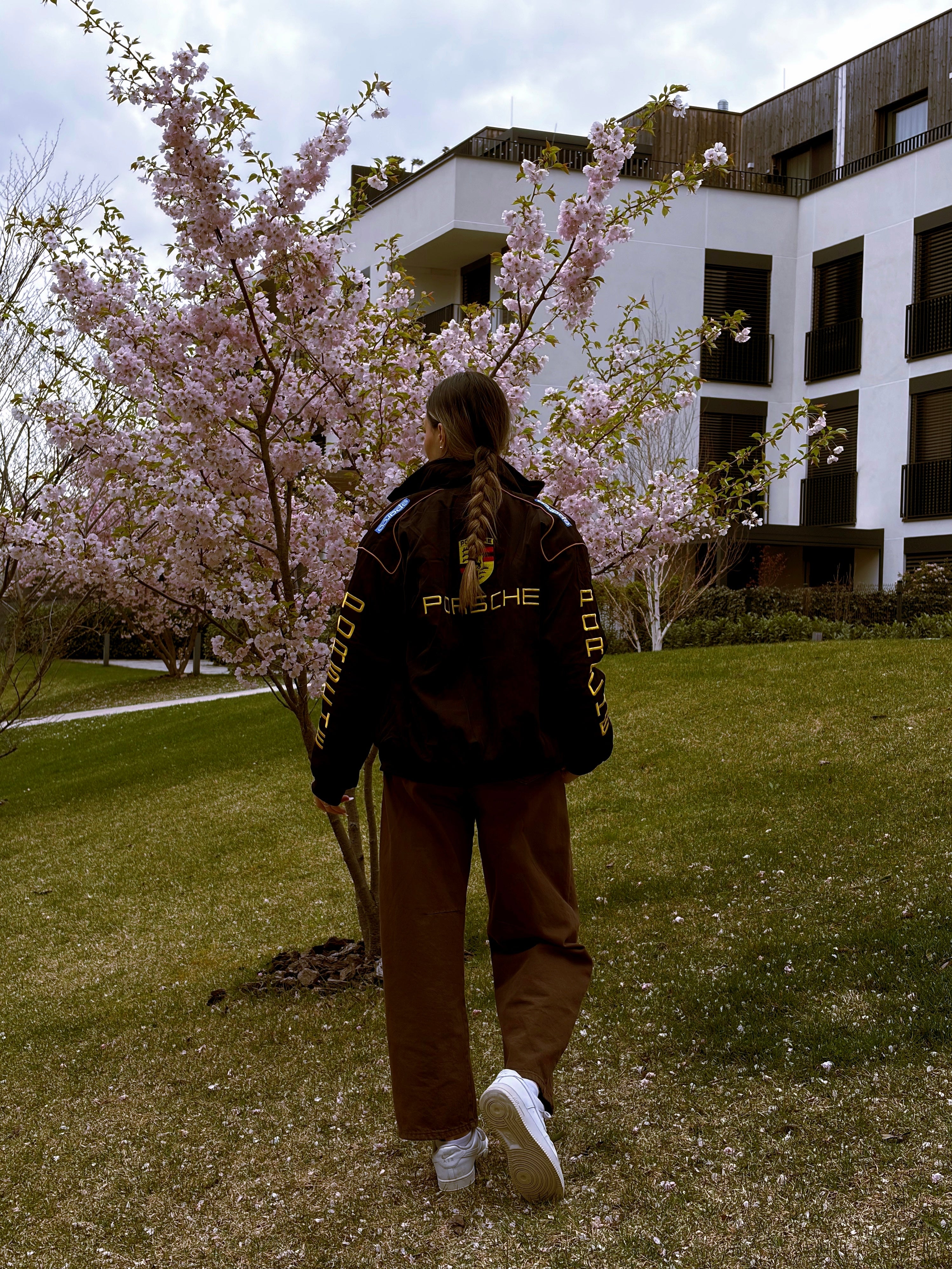 A person with a braid, dressed in the Porsche Vintage Racing Jacket and brown pants, walks on grass near a blooming cherry blossom tree, with a modern building in the background—a stylish nod to motorsport heritage that seamlessly blends into the vibrant spring scene.