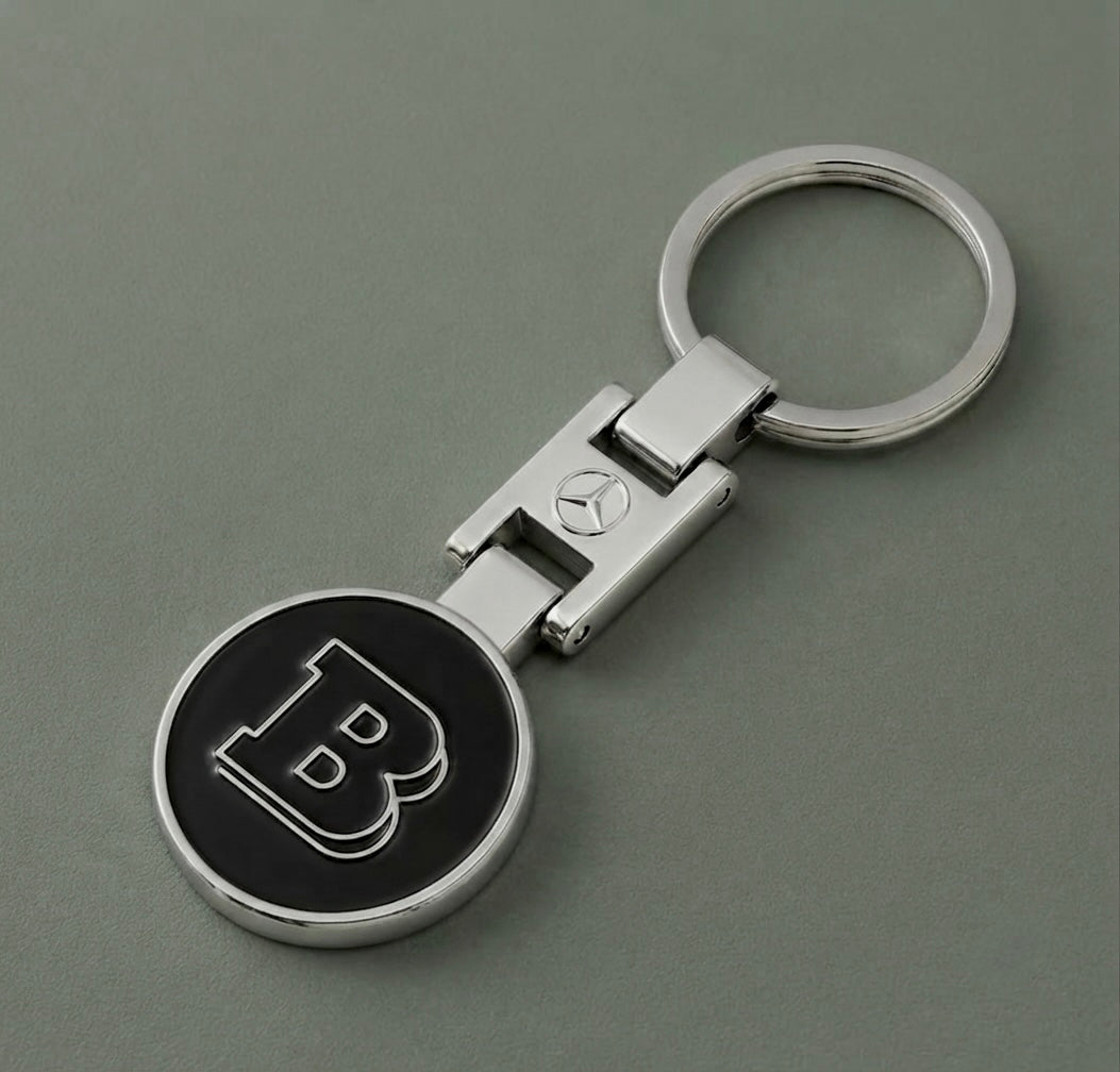 The Brabus Keychain features a polished silver finish with a round black emblem displaying the iconic bold “B” and includes a small Mercedes-Benz logo on the connector. Displayed on a sleek gray surface, it’s an ideal accessory for performance enthusiasts and Mercedes-Benz tuners eager to express their passion for high-performance cars.