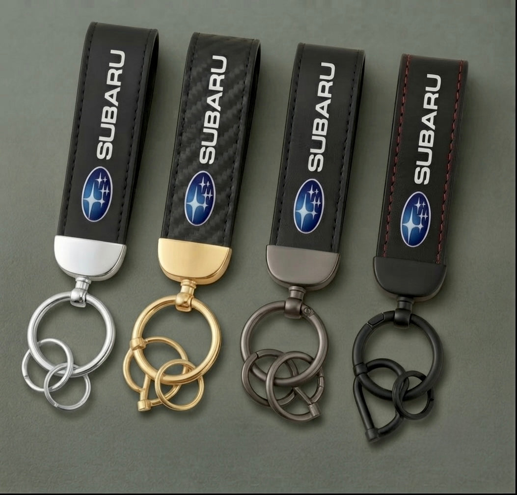 Four Subaru Keychains, each with a distinct metal finish—silver, gold, gray, and black—are arranged side by side on a dark surface. Each keychain features the Subaru logo and brand name on a black strap. These stylish Subaru Keychains make an elegant and functional accessory for enthusiasts looking to enhance their everyday carry.