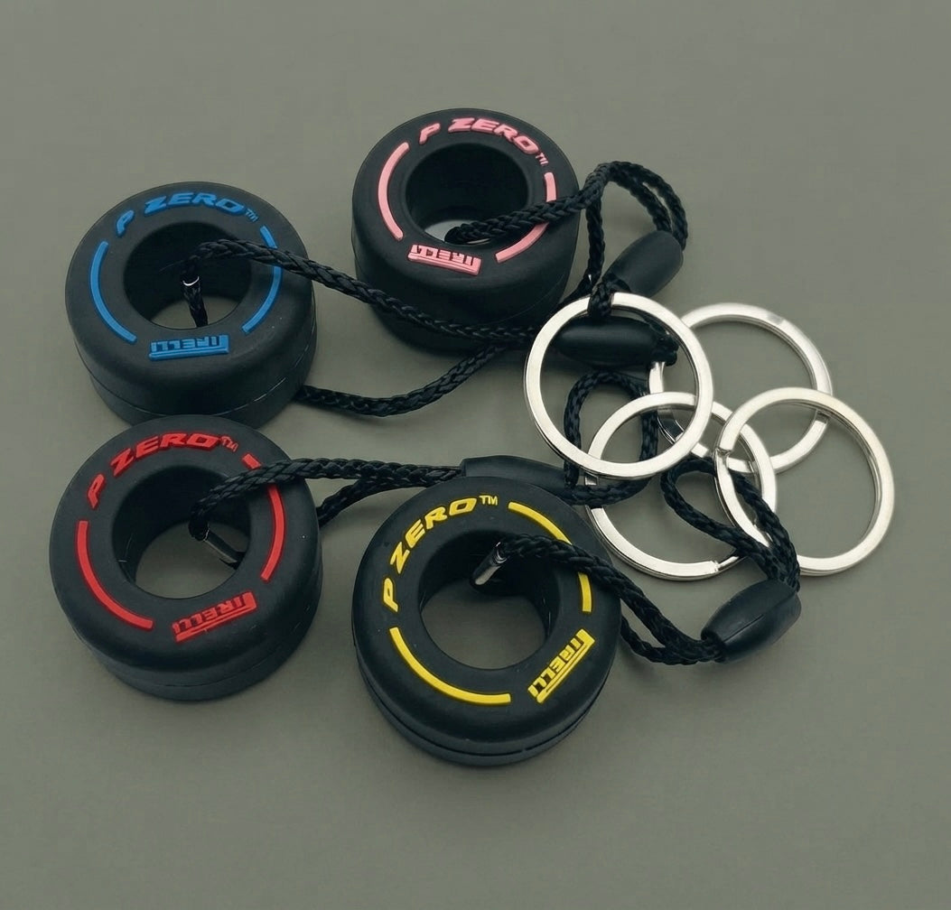 Four Pirelli Tire Keychains—each designed to resemble miniature Pirelli P Zero tires with blue, pink, and yellow accents—are attached to black cords and silver key rings, making them a perfect Formula 1 F1 fan gift. The keychains are arranged on a gray background.