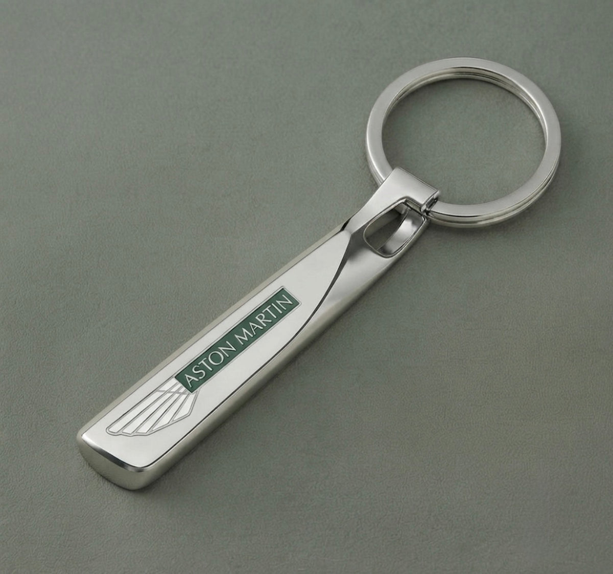 The Aston Martin Keychain features a polished silver finish and a rectangular tag showcasing the iconic green and white Aston Martin logo with wings, set on a grey surface—an essential luxury accessory for any car enthusiast.