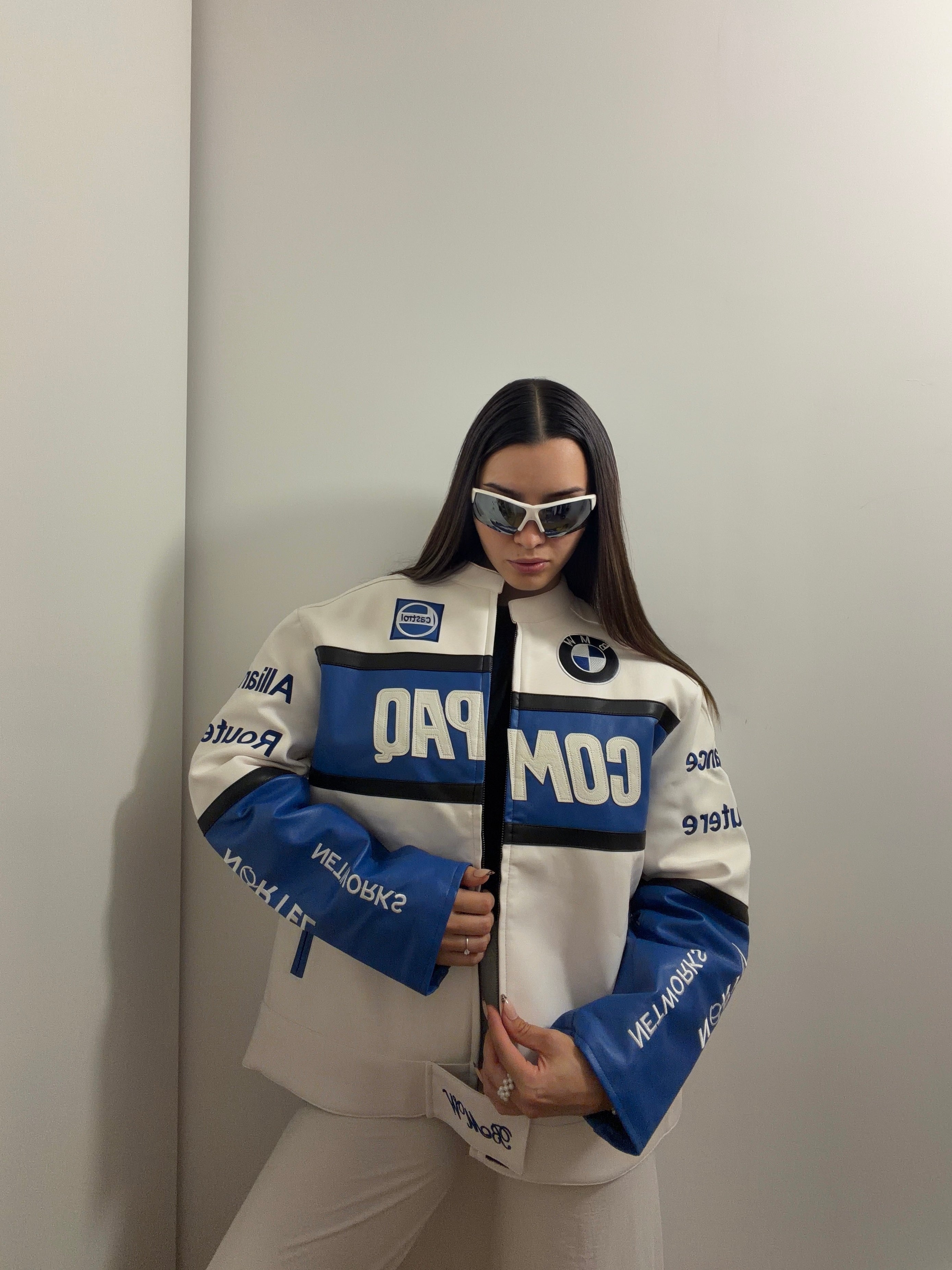 Clad in the BMW Vintage Leather Jacket, distinguished by its racing-inspired design and prominent COMPAD text, an individual wearing futuristic sunglasses strikes a confident pose against a plain, light backdrop—an effortless embodiment of motorsport heritage.