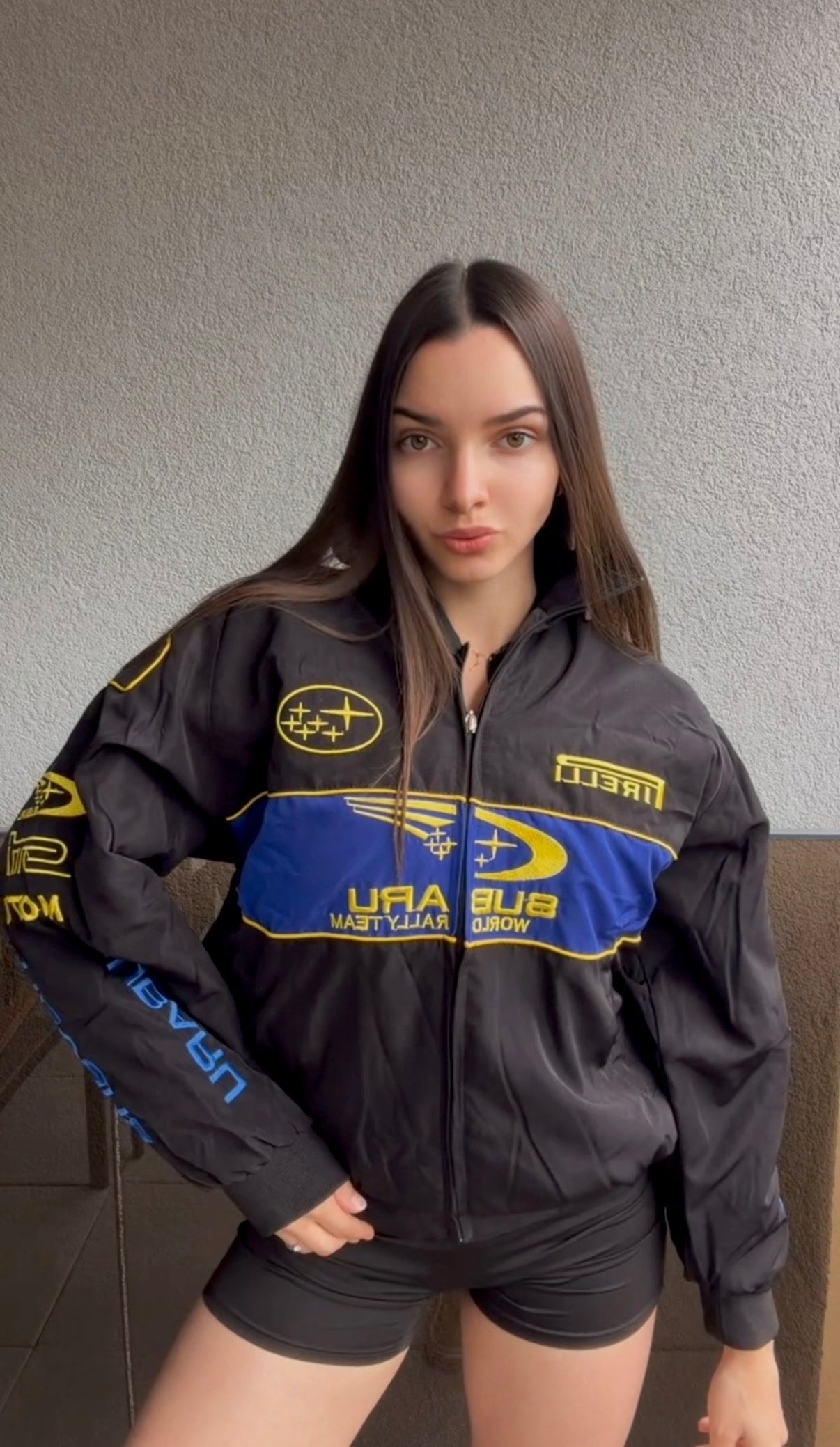 A person with long brown hair stands indoors, wearing the Subaru Rally Vintage Racing Jacket featuring blue and yellow Subaru World Rally Team logos, looking directly at the camera with a neutral expression.